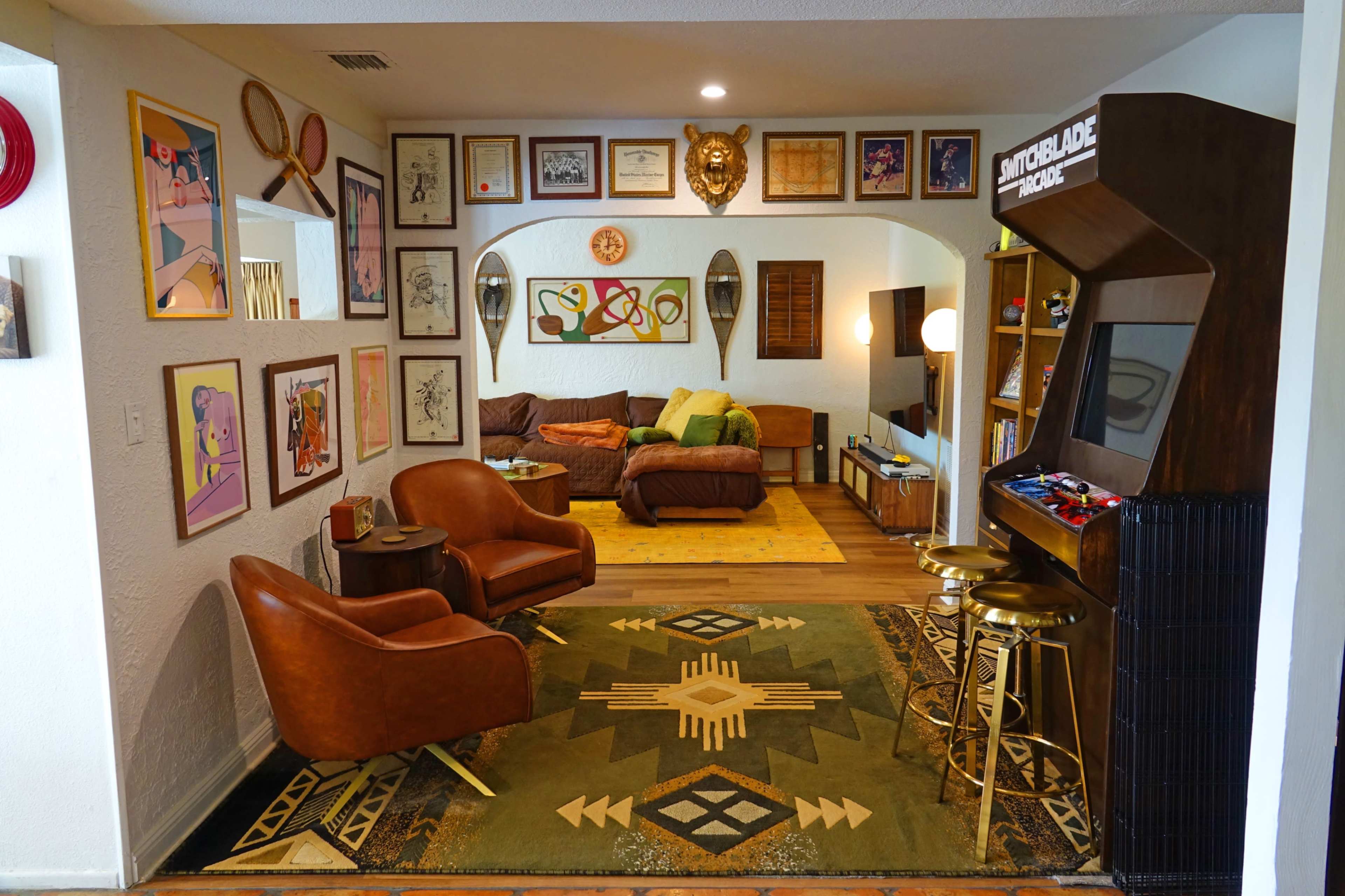 The image shows a mid-century style living room featuring two brown armchairs, a large rug with geometric patterns, and an arcade cabinet against the wall amid framed artwork and a cozy sofa area.