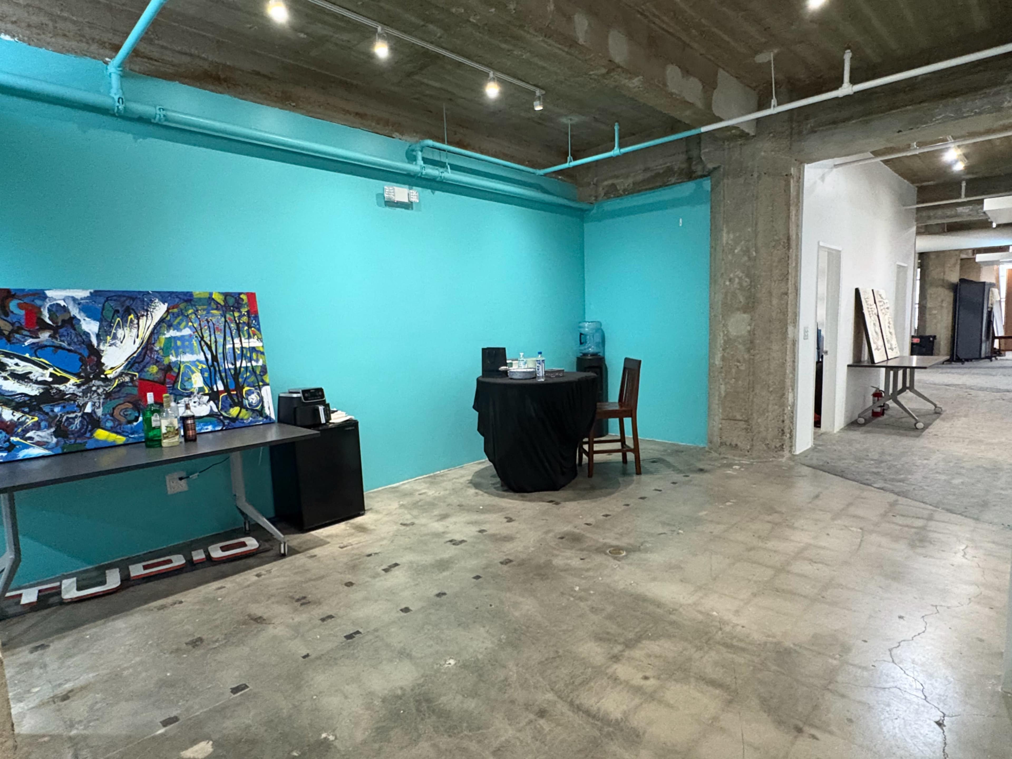 The image shows an open, minimalist studio space with a turquoise wall, a black table with a chair, and abstract artwork hanging nearby.