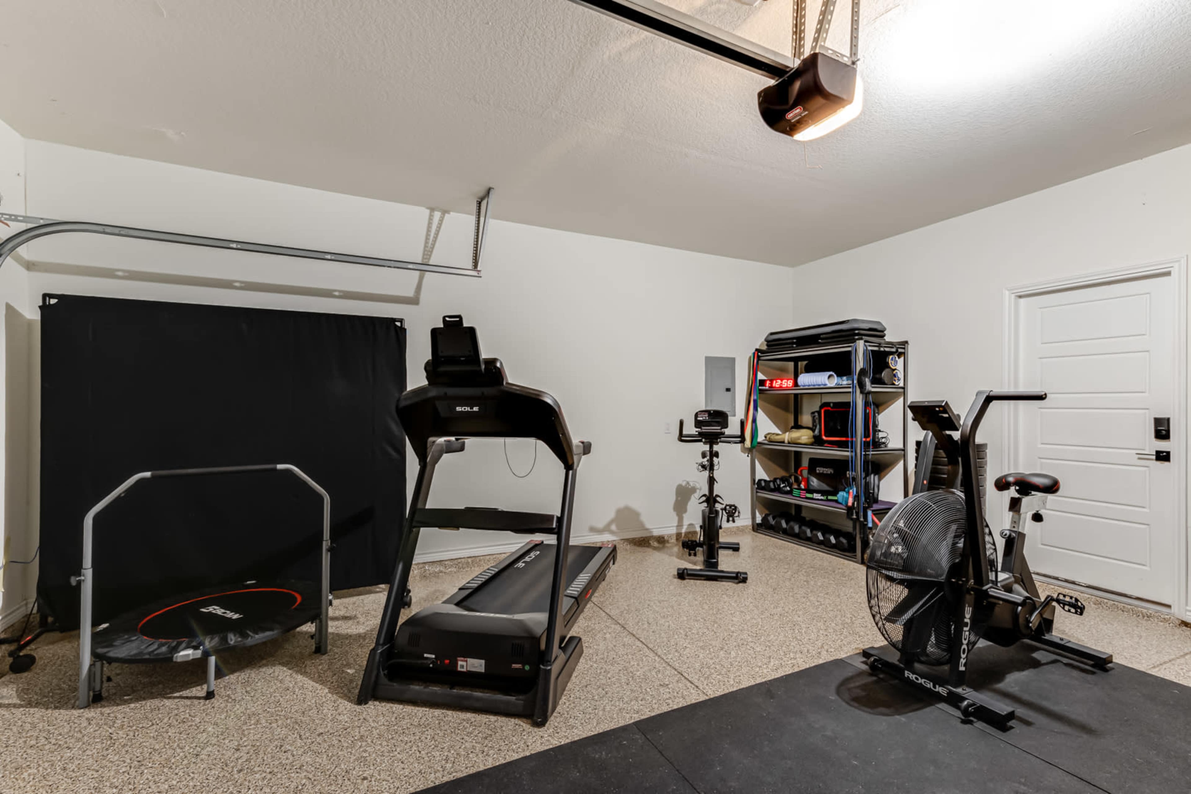 A home garage equipped with a treadmill, a stationary bike, a trampoline, and various fitness equipment arranged on a shelf.