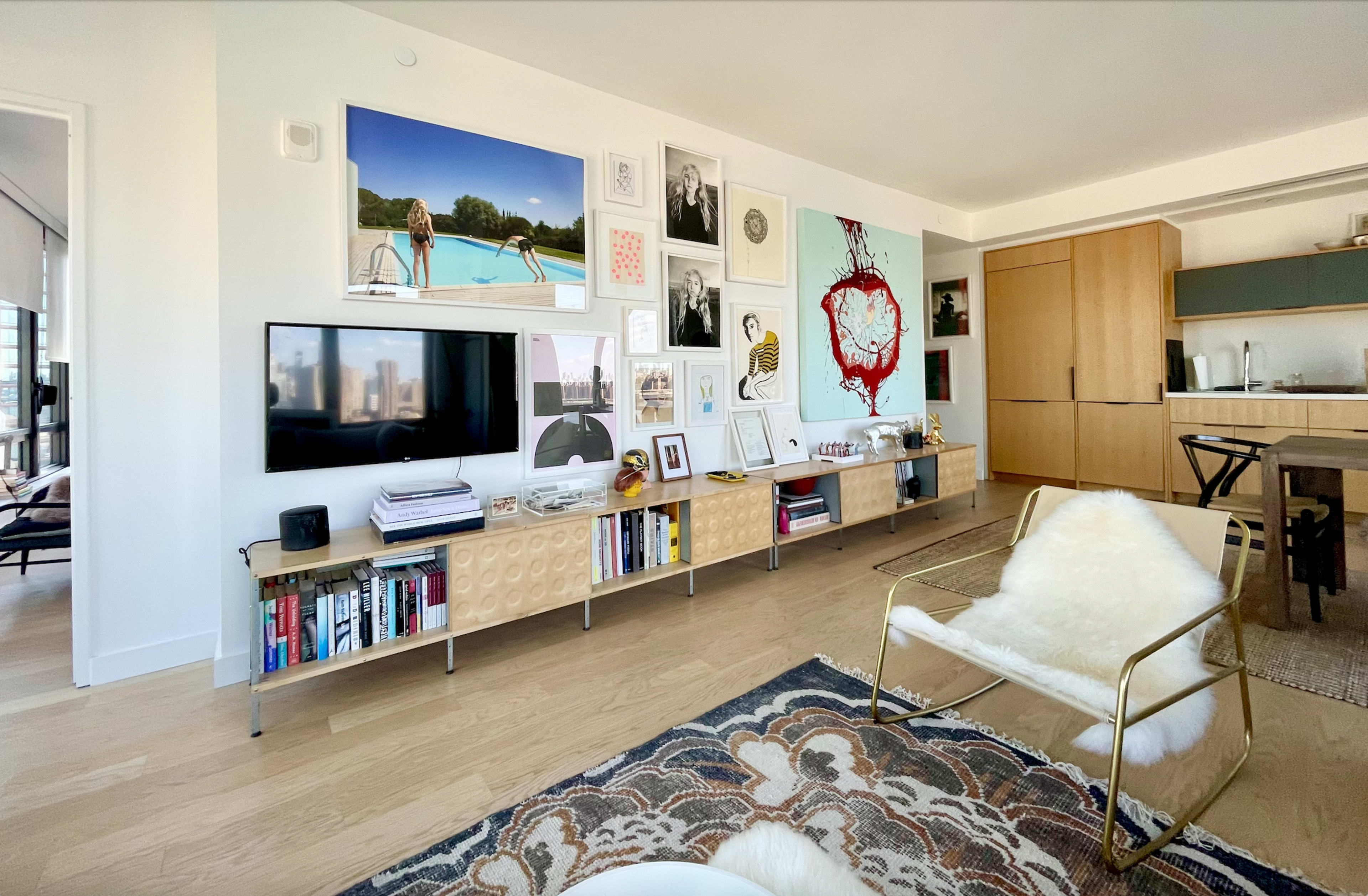 The image depicts a modern living area featuring a wall-mounted television, a collection of framed artworks, and a wooden shelving unit filled with books and decorative items.