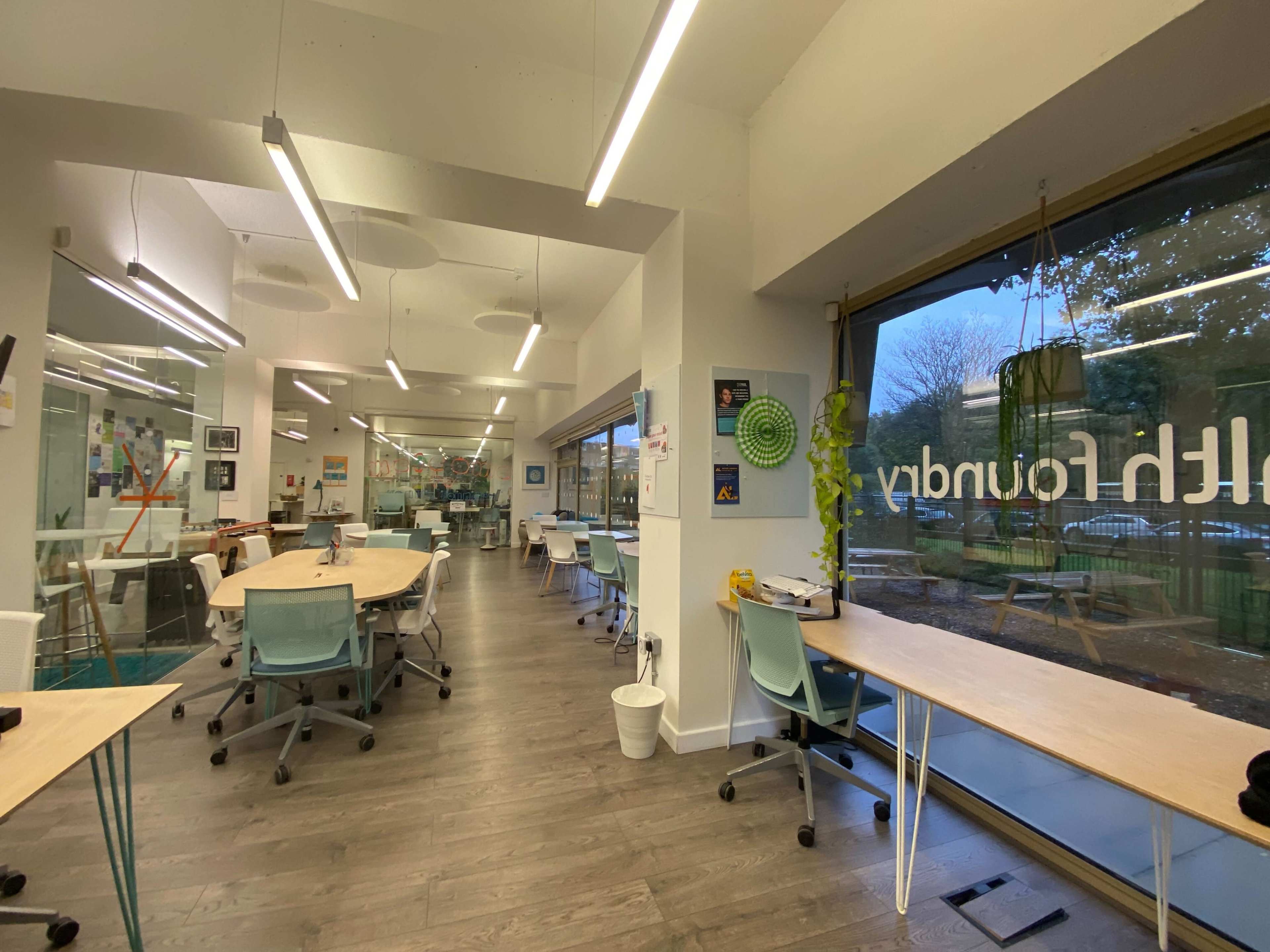 The image shows a modern, open workspace with several tables and chairs arranged in a well-lit room featuring large windows.