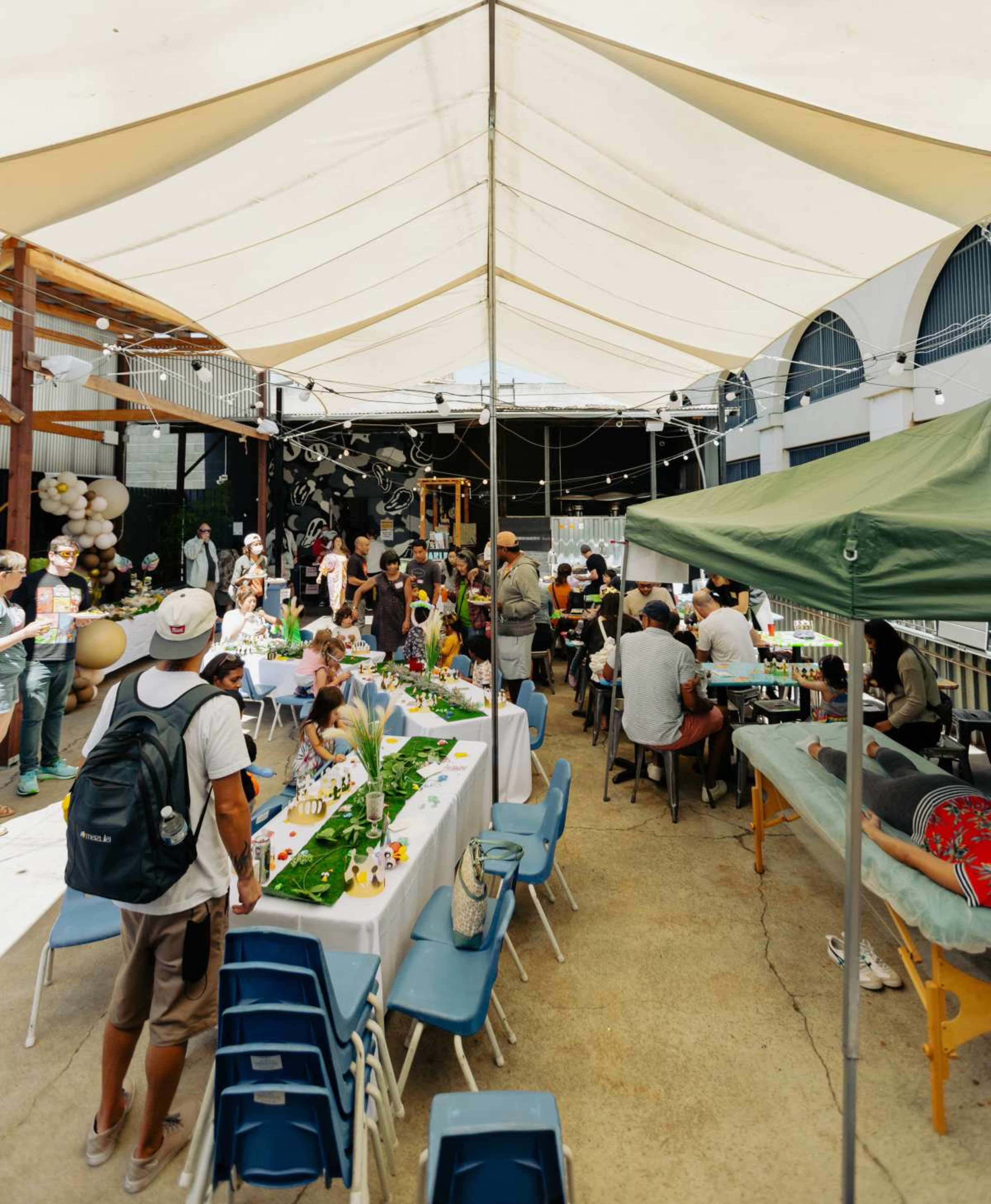 A large outdoor gathering is taking place under a tent, with tables set up for dining and decorations visible in the background.
