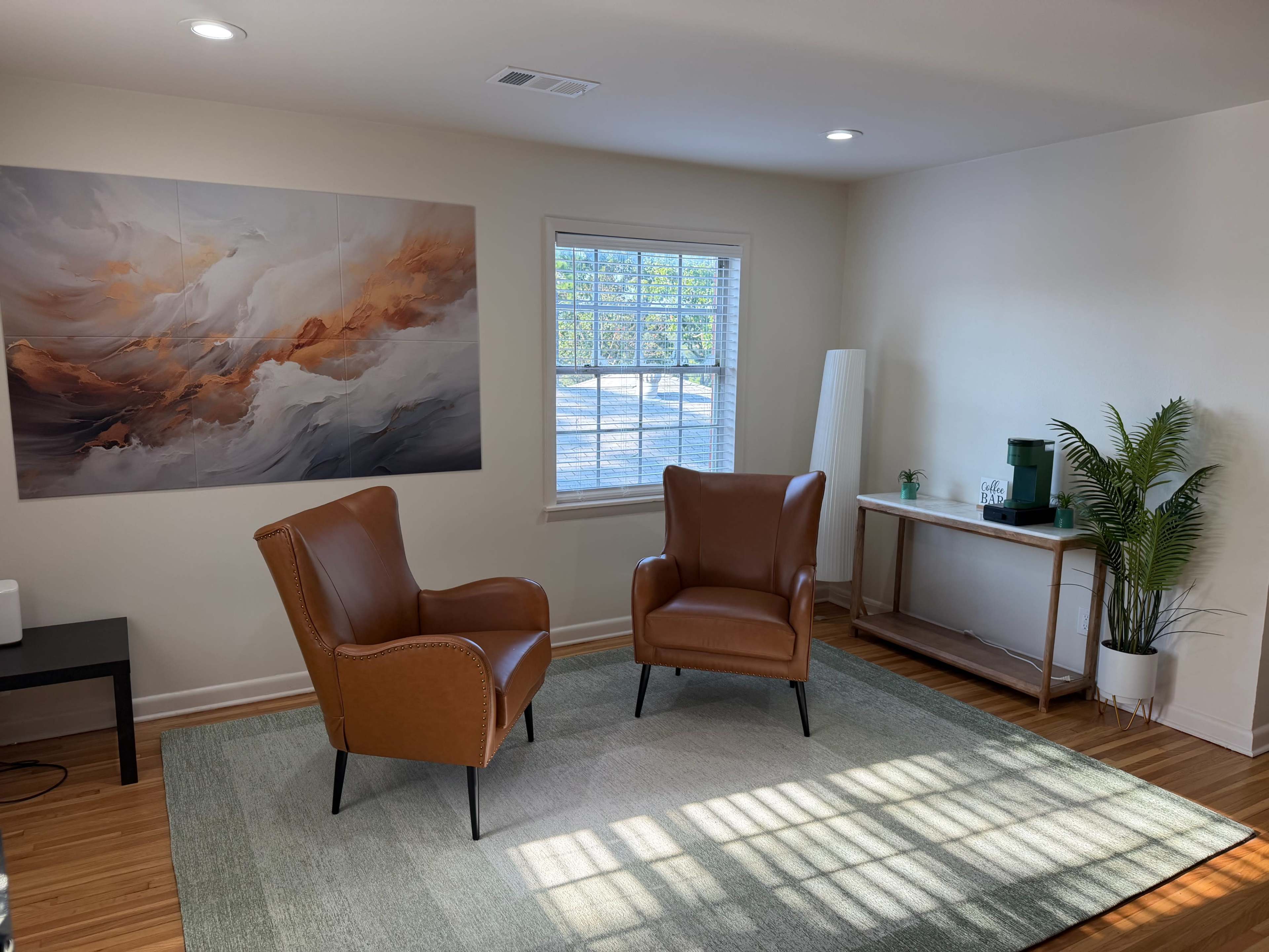 The image shows a bright room featuring two brown leather chairs positioned on a rug, with a large abstract painting on the wall and a small table with a plant in the corner.