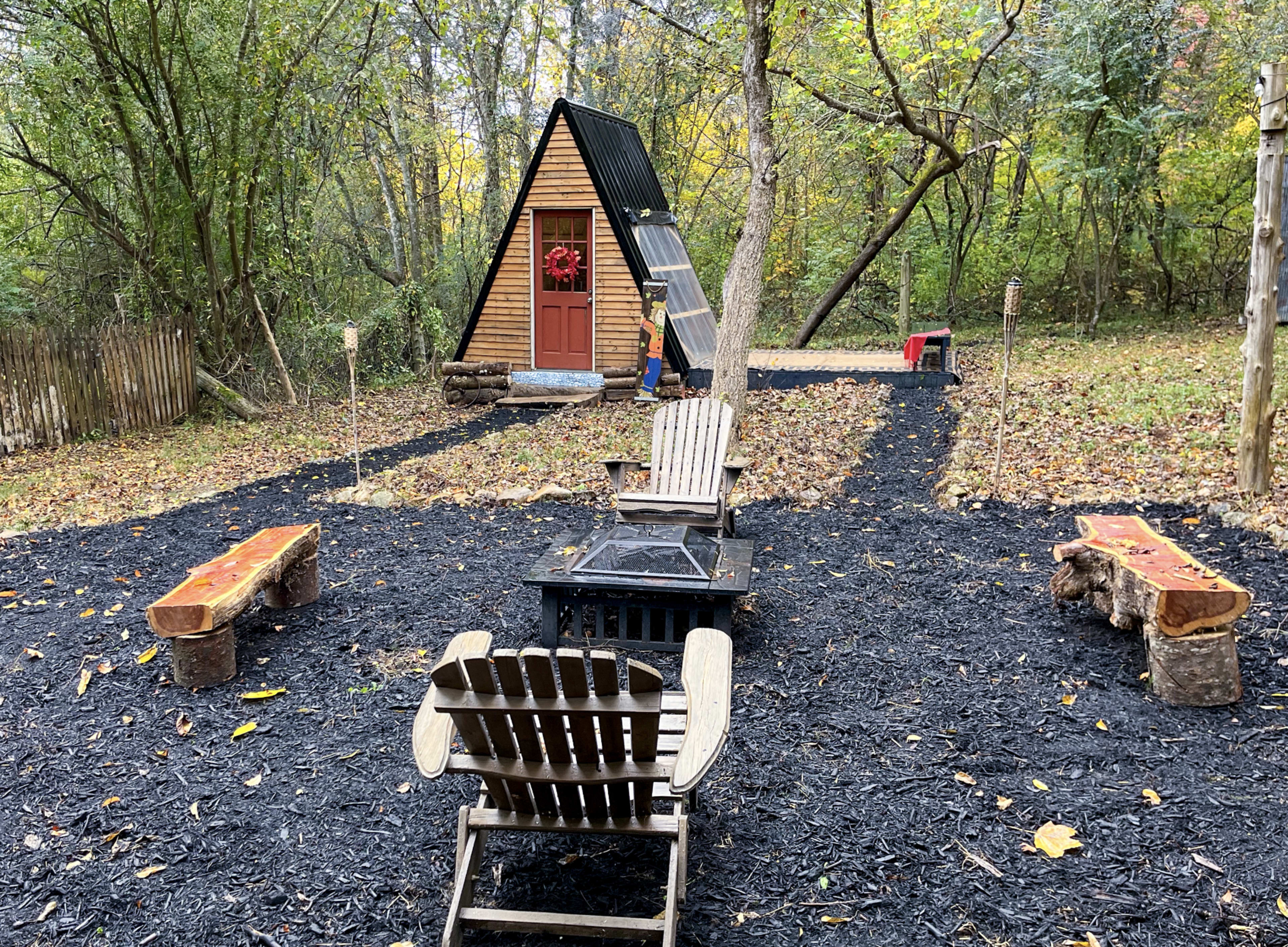 A small A-frame cabin is surrounded by wooden benches and a fire pit, set amidst a wooded area with autumn foliage.