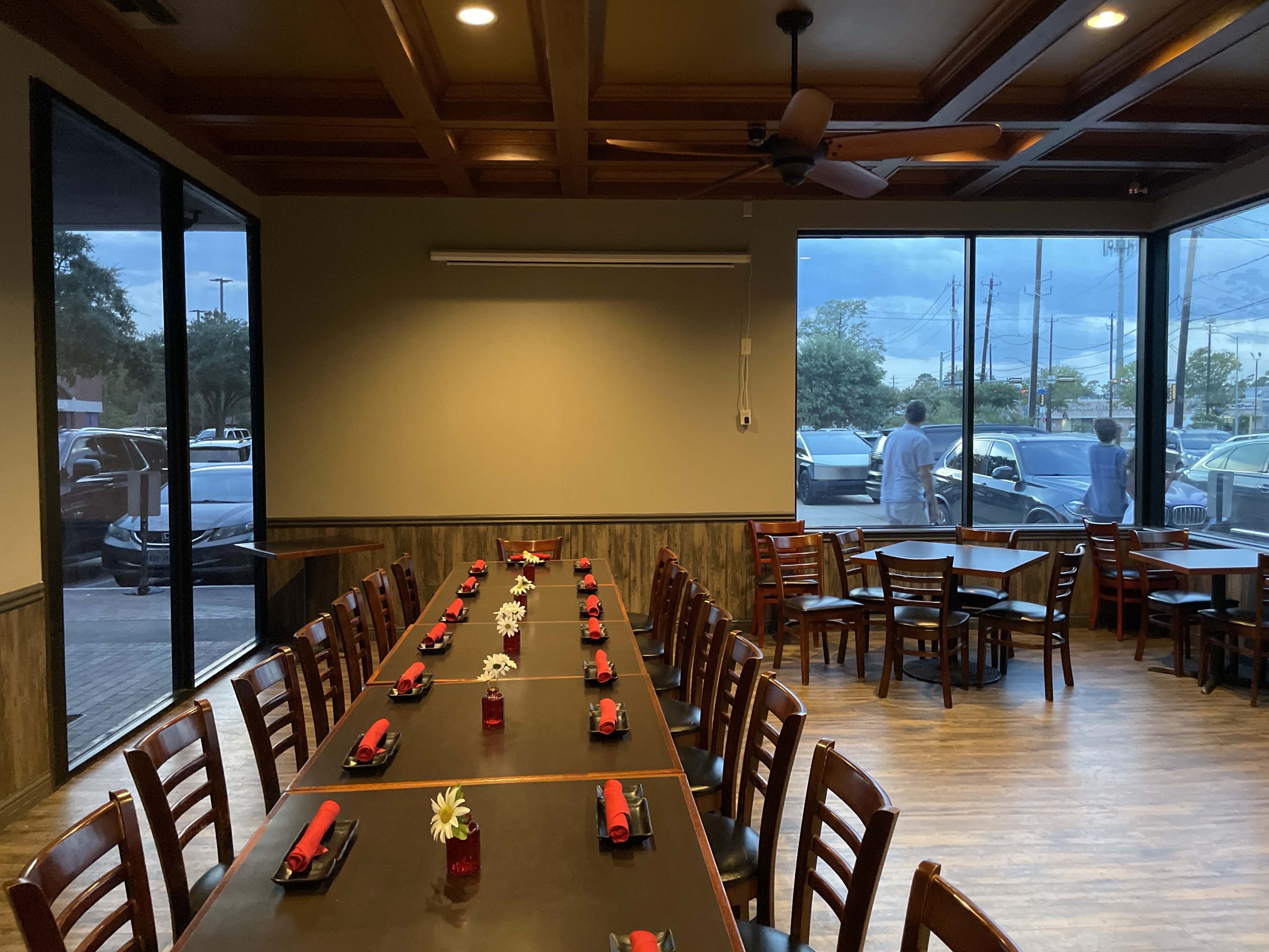 A long table is set for dining in a restaurant, with red napkins and small flower centerpieces, while two people stand outside looking in through large windows.