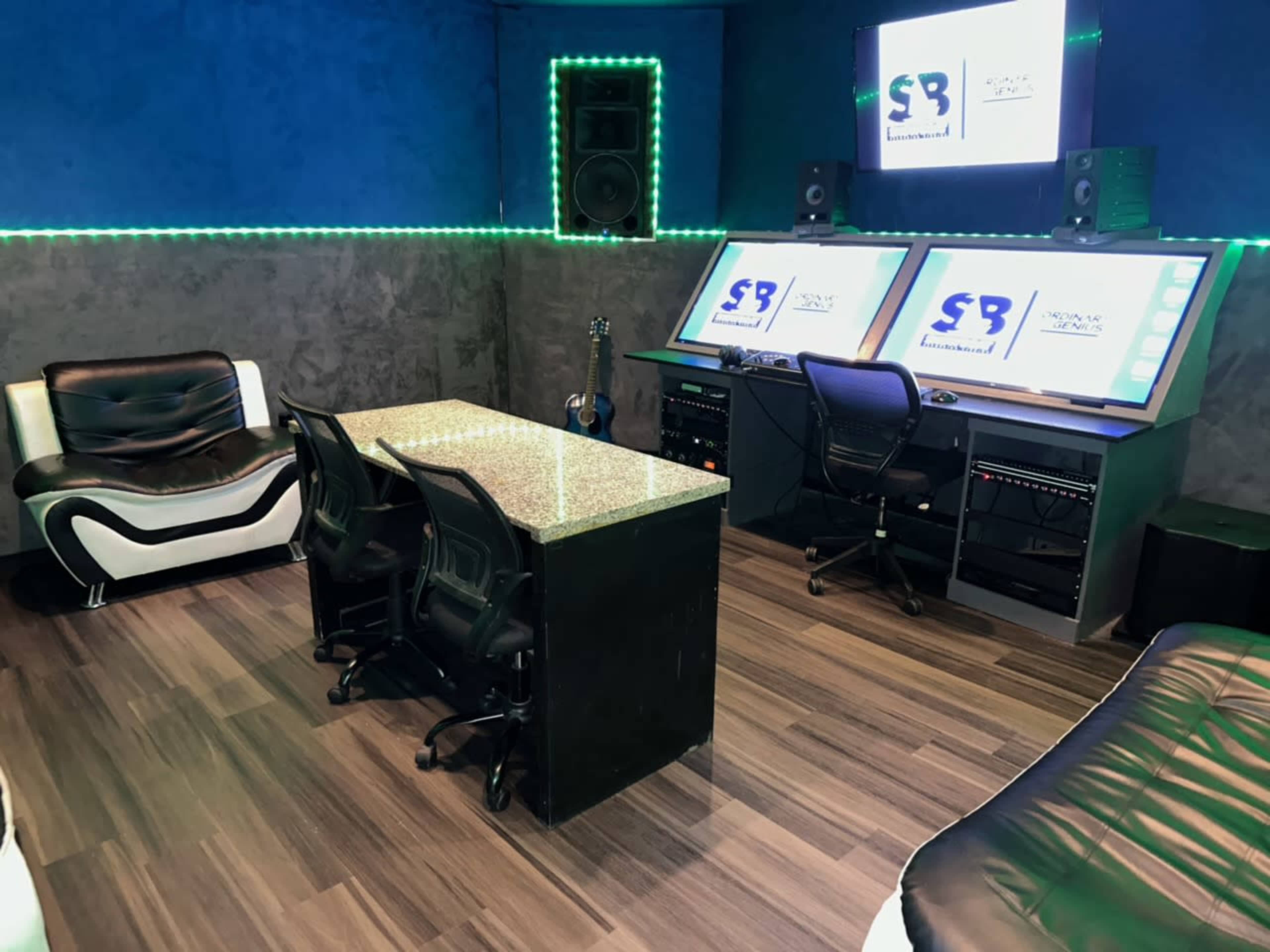 The image shows a modern recording studio with two large mixing consoles, a glass-topped table, black chairs, and blue wall accents illuminated by green LED lighting.