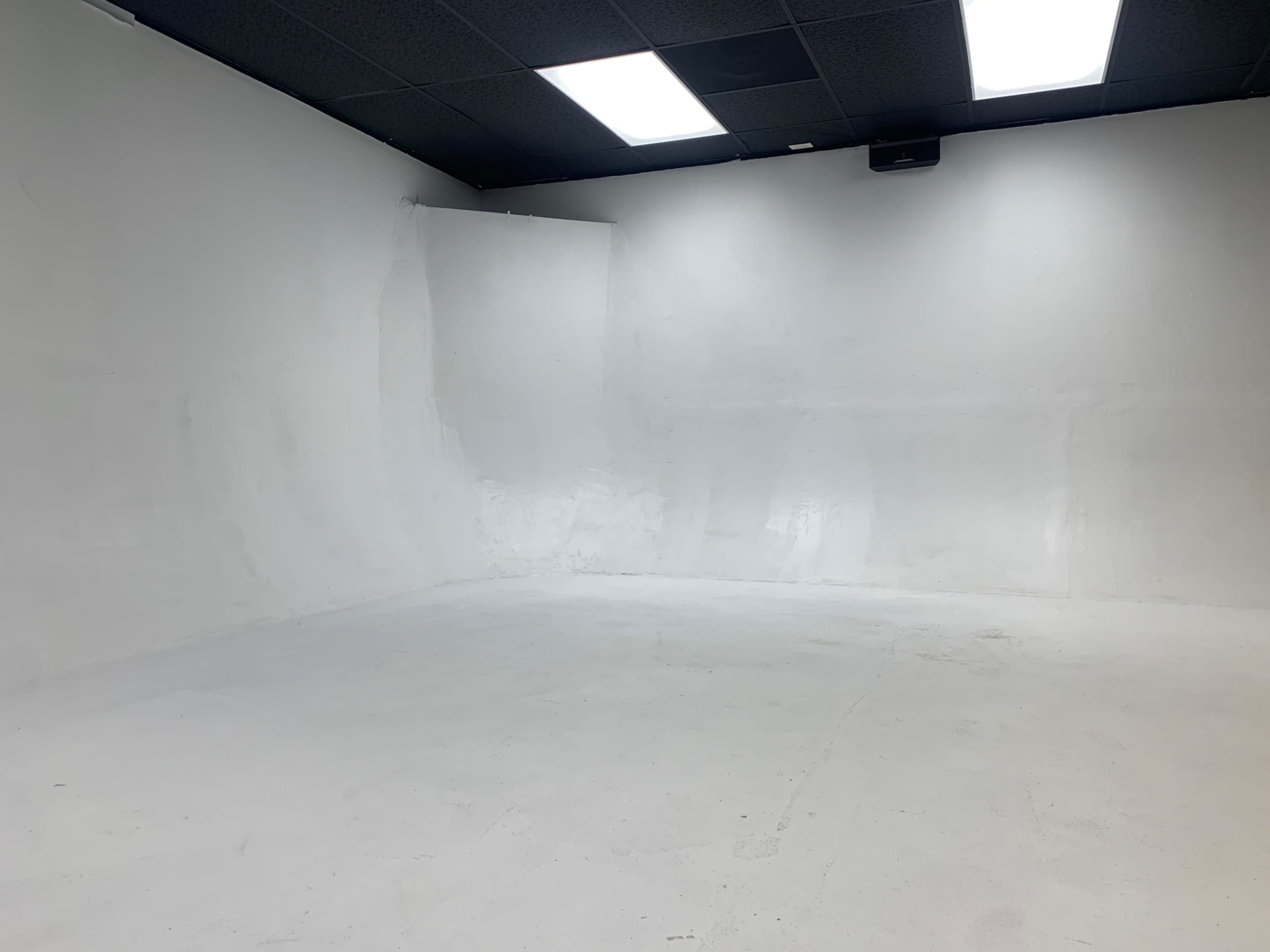 A large, empty room with white walls and a smooth, white floor, illuminated by fluorescent lights.