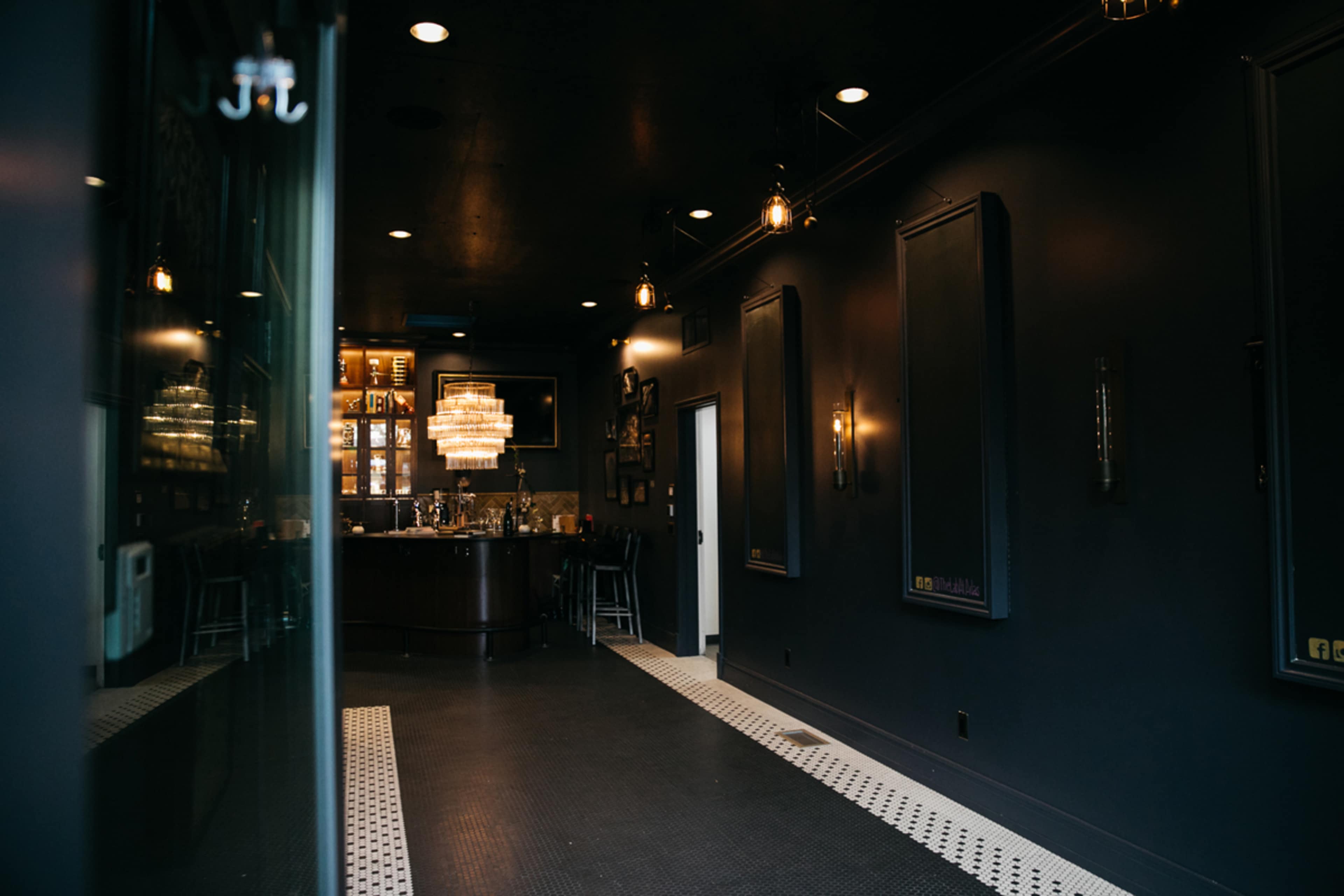 A dimly lit bar area with dark walls, a chandelier, and a sleek counter visible at the end of a hallway.