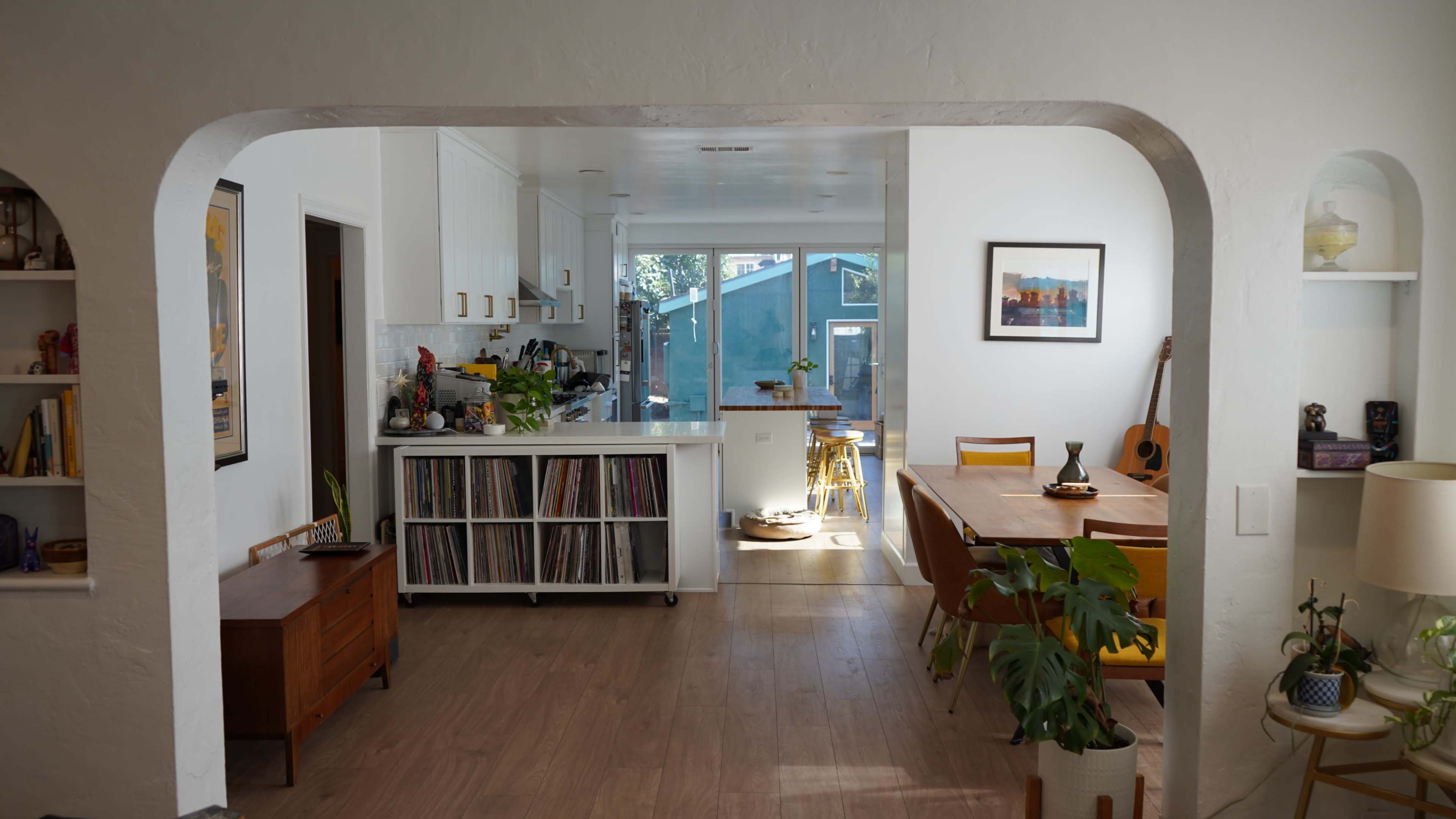 A bright, open living space features a kitchen area with a vinyl record shelf, a dining table, and large windows that bring in natural light.