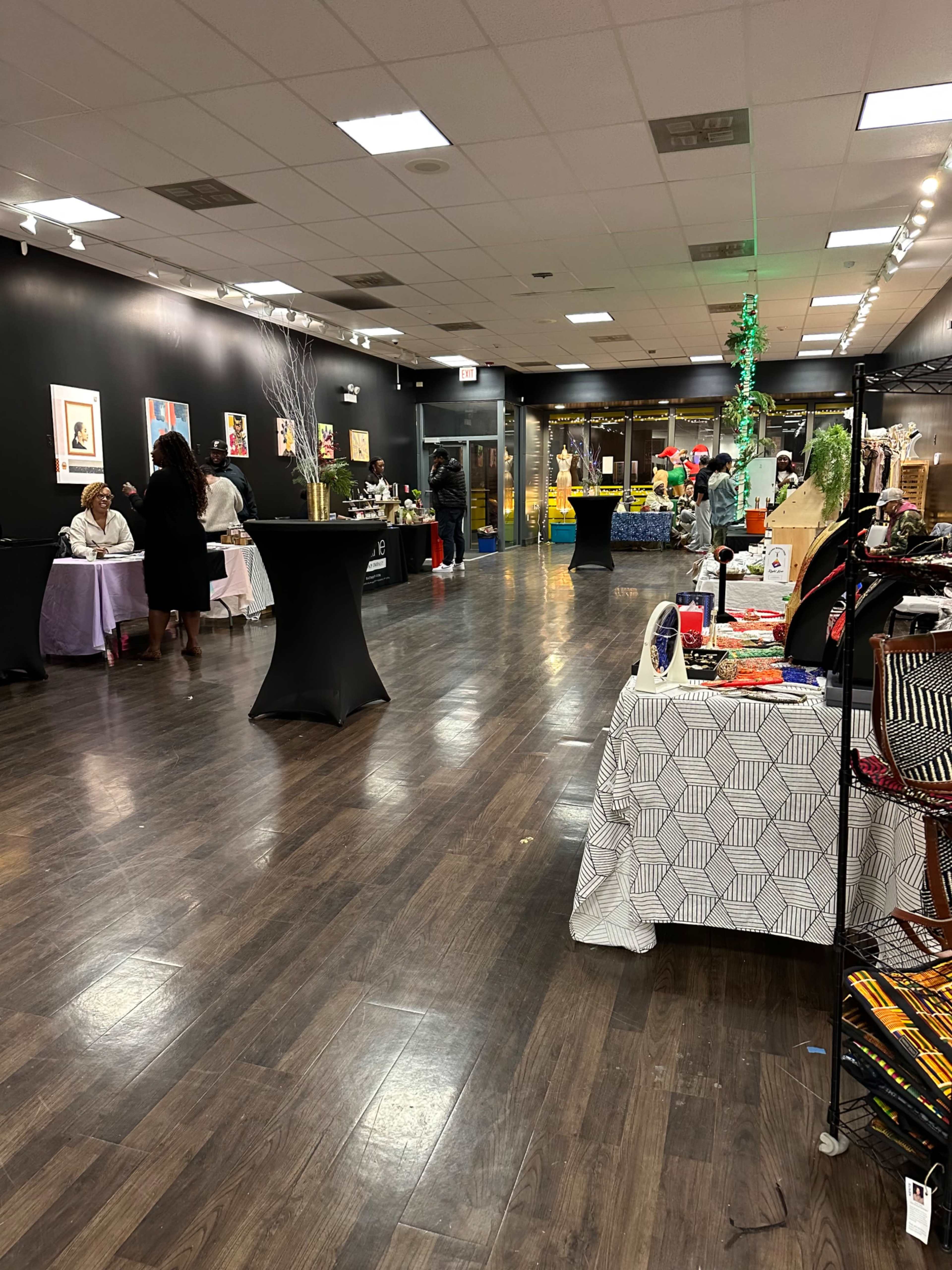 The image shows an indoor event space with various tables displaying artworks and crafts, surrounded by attendees engaging in conversation.
