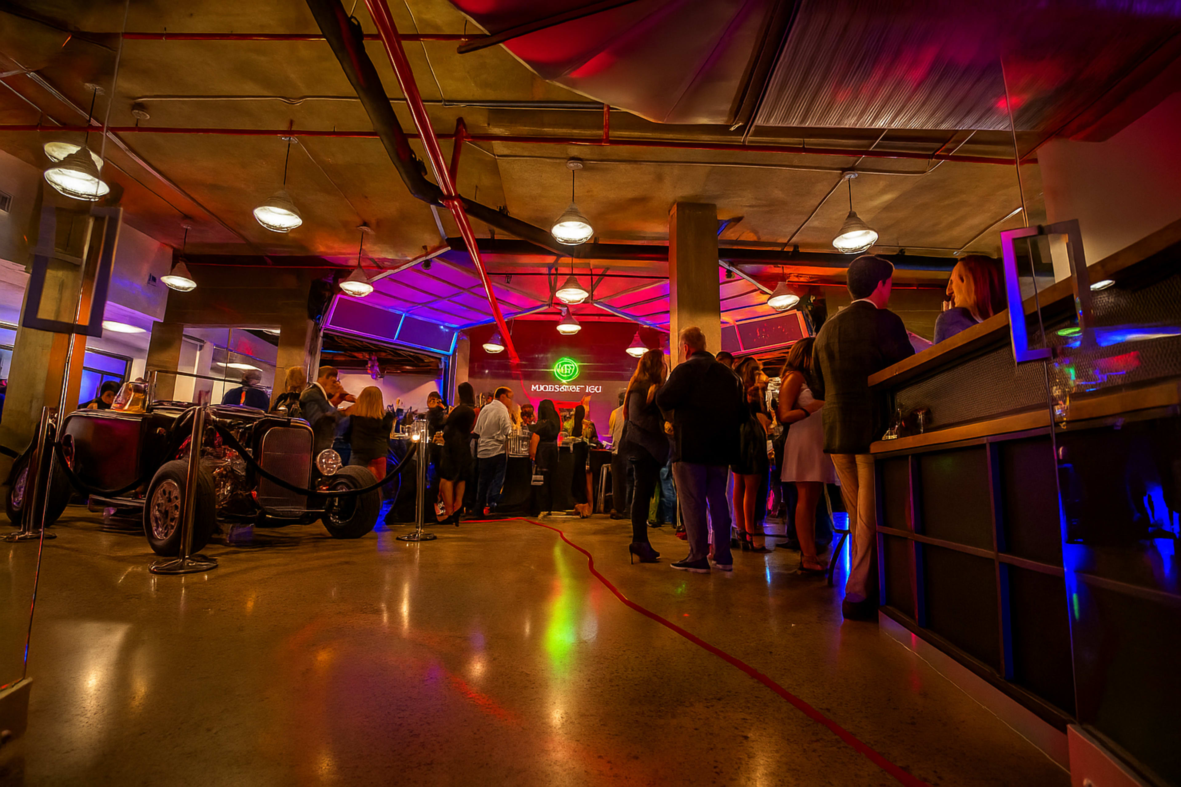 A crowd gathers in a dimly lit event space featuring colorful lighting and a vintage car display.