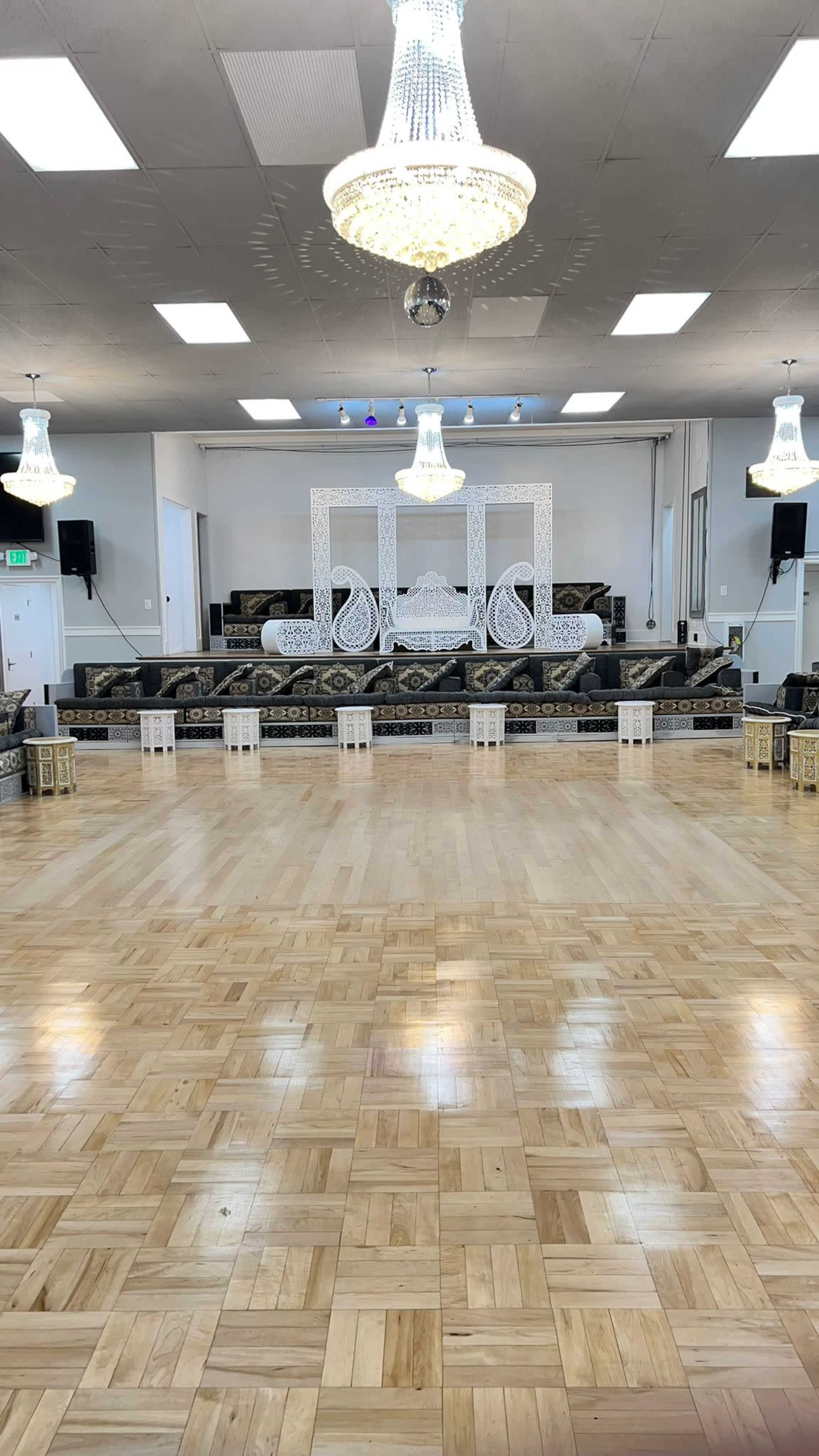 An empty hall with a wooden floor, adorned with chandeliers, featuring a decorative stage at one end.