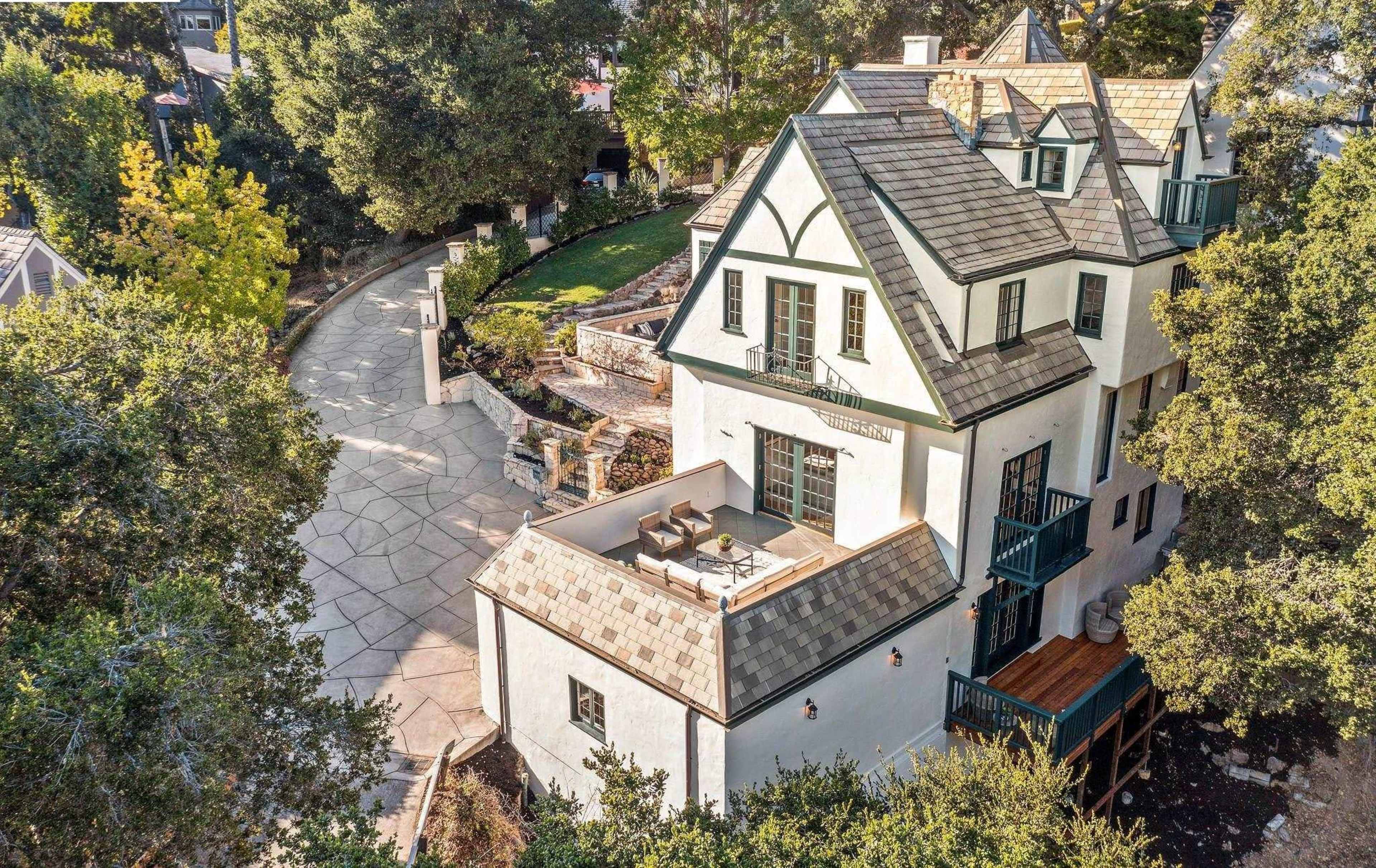 A large, three-story house with a steeply pitched roof and balcony is situated amidst trees, with a winding driveway leading up to it.