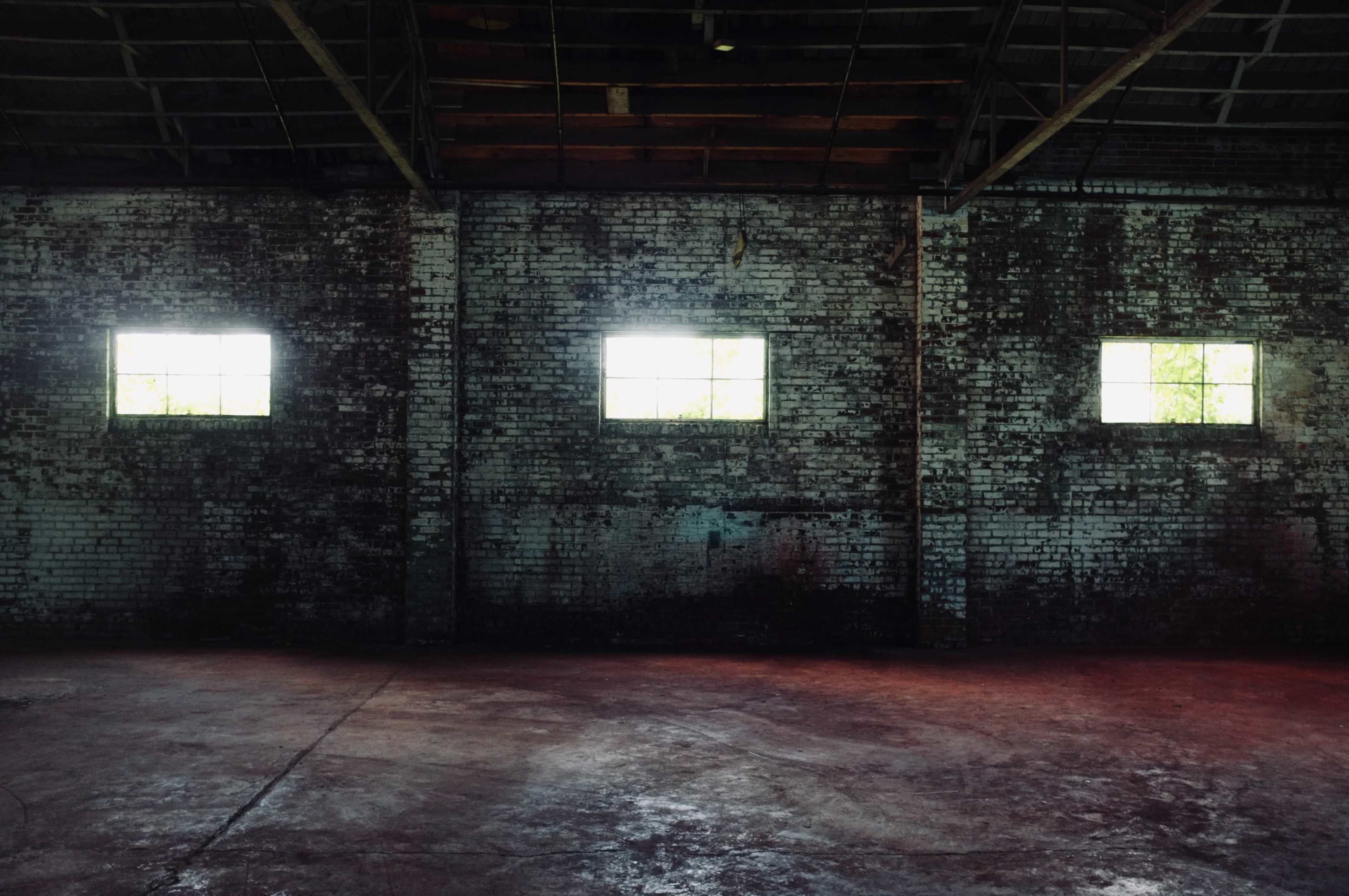 The image shows an empty industrial space with four small windows in the brick walls, allowing light to filter in.