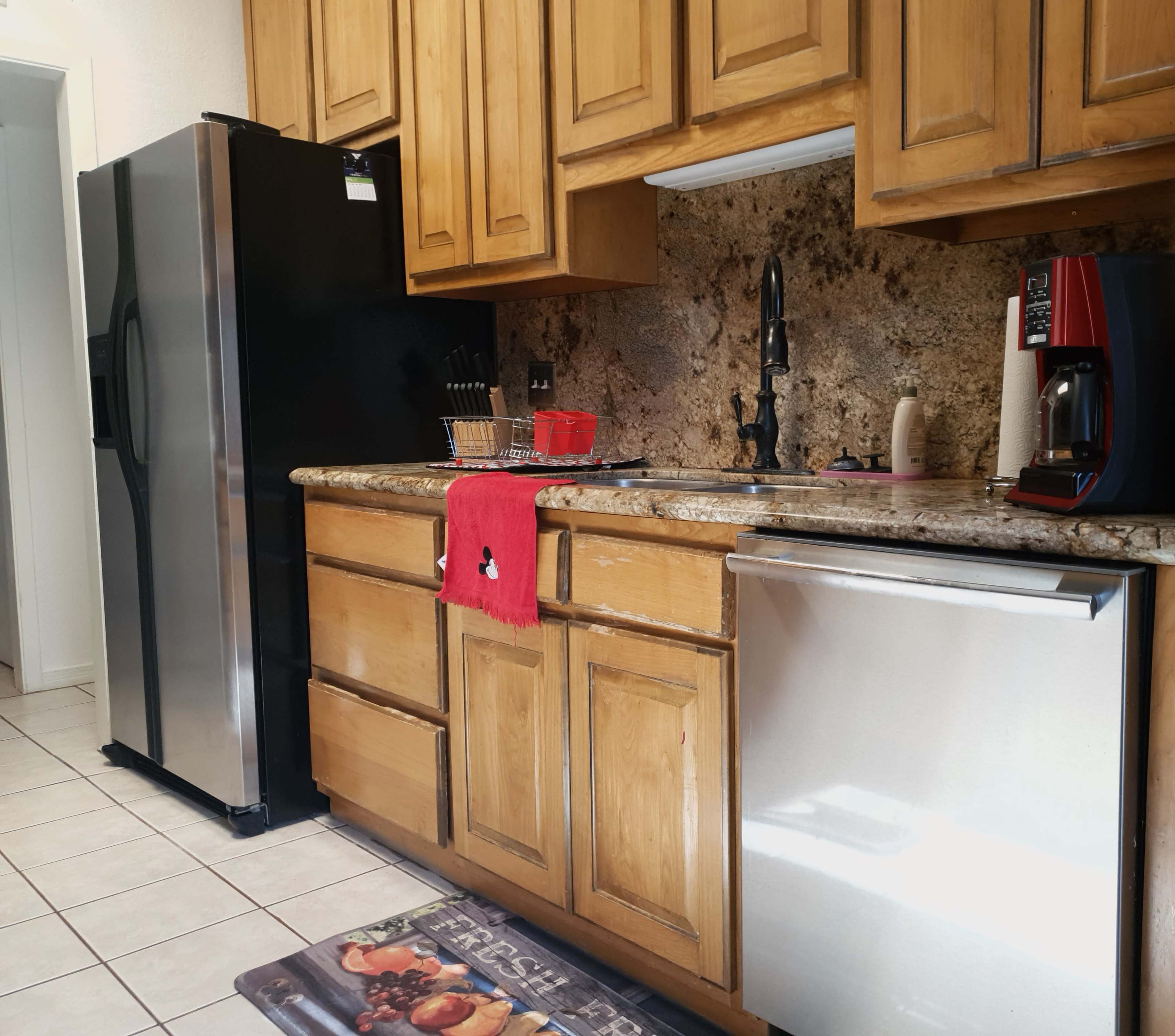 The kitchen features wooden cabinets, a stainless steel refrigerator, a coffee maker, and a dishwashing machine, all arranged along a tiled floor.