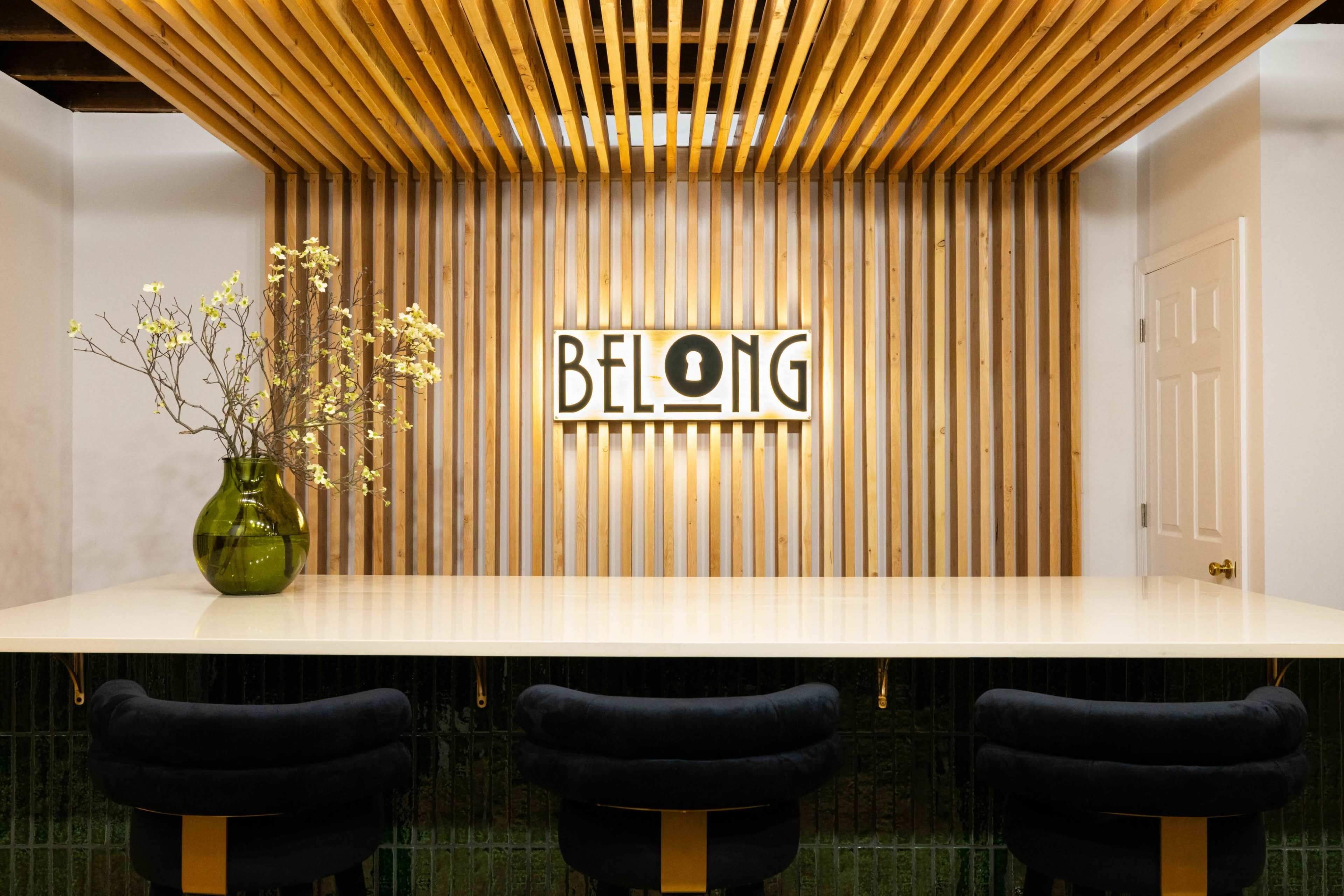 A modern interior with a wooden slatted wall featuring a sign that says "BELONG" above a sleek bar with dark cushioned stools and a green vase holding flowers.