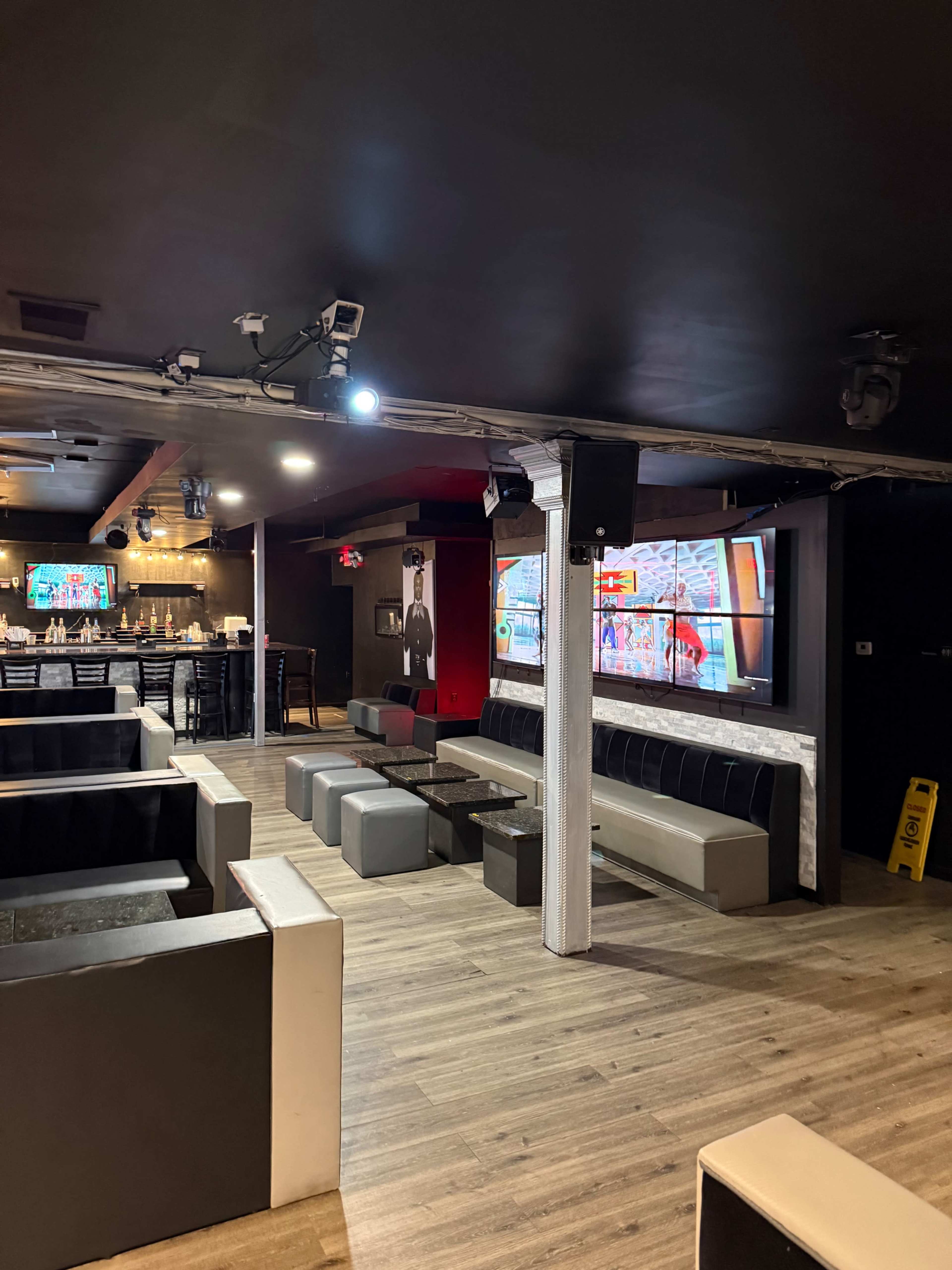 The image shows a modern lounge area with black and gray seating, multiple screens displaying content, and a wooden floor.