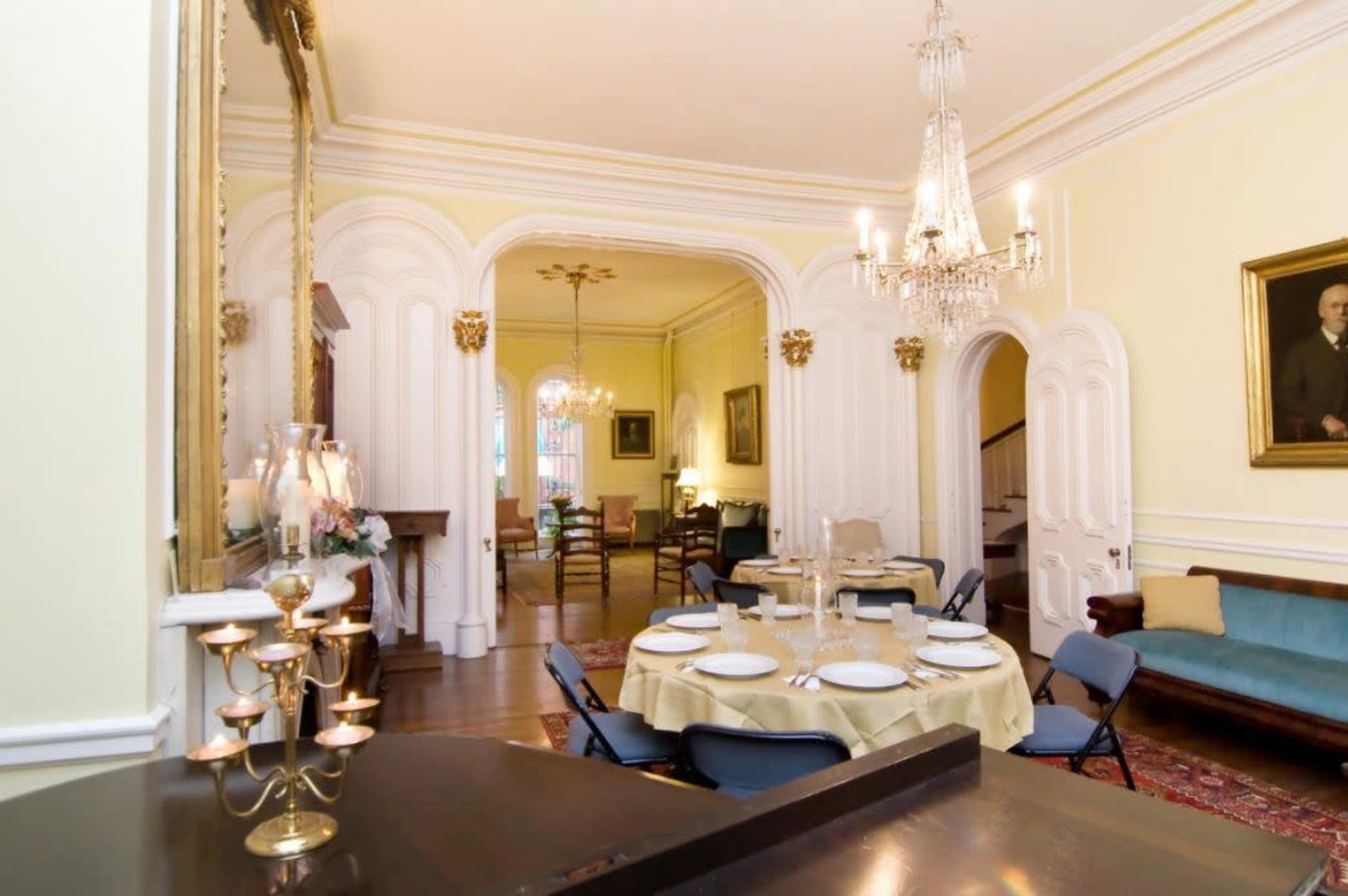 A formal dining room features a round table set with dishes, surrounded by chairs, and is adorned with a chandelier and elegant decor.