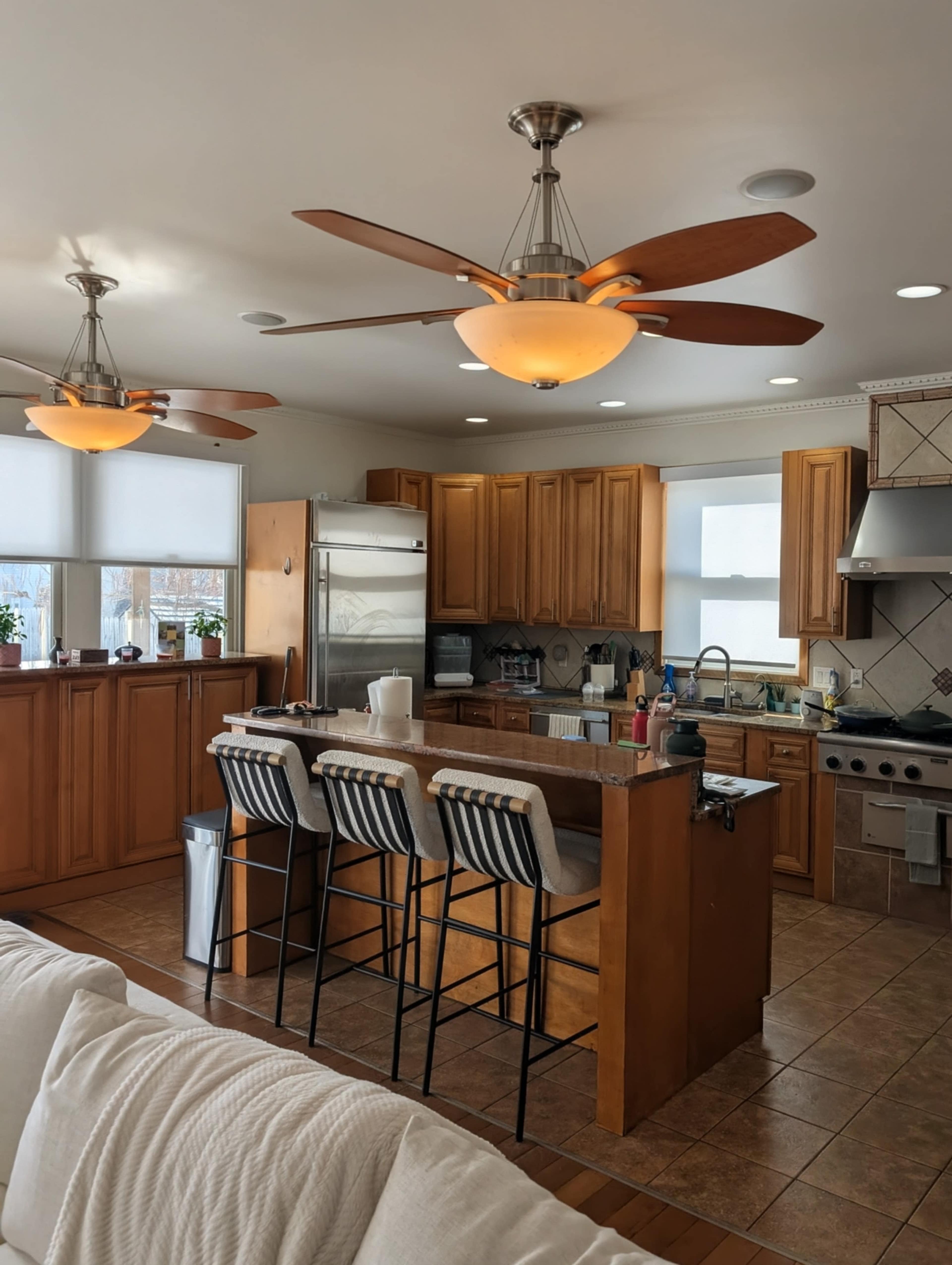 A modern kitchen features wooden cabinetry, an island with bar stools, stainless steel appliances, and two ceiling fans with lights.