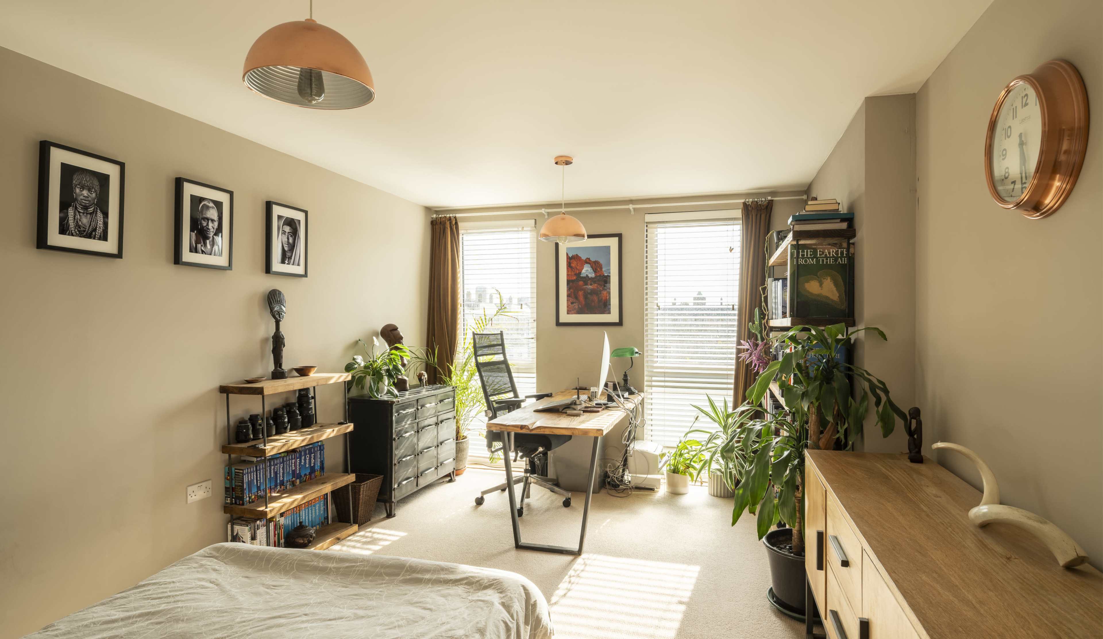 The image shows a well-lit room with a workspace featuring a desk, chair, and plants, surrounded by framed photographs on the walls.