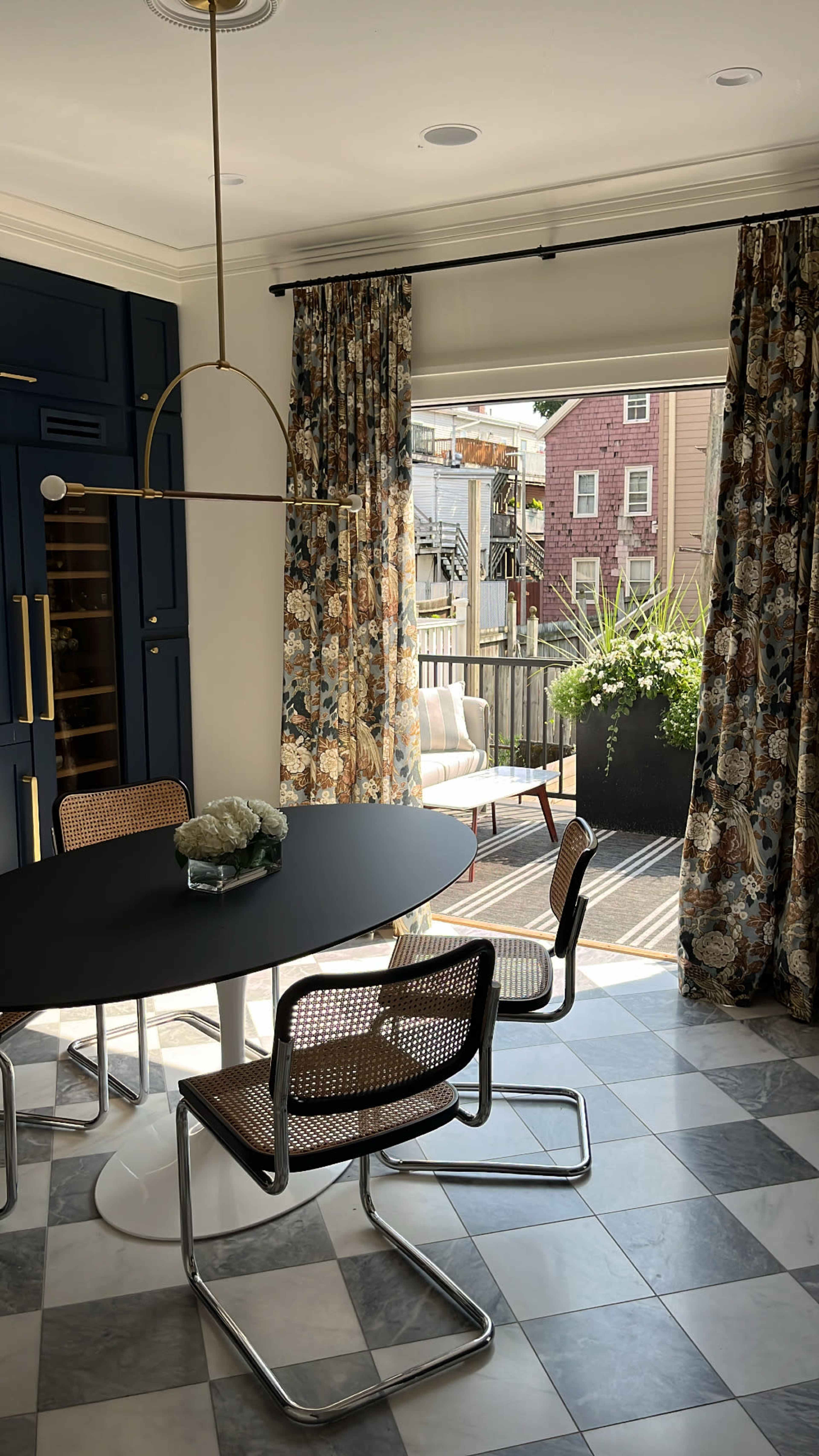 A round black dining table with four chairs is set in a brightly lit room featuring patterned curtains and a door leading to a deck with potted plants.
