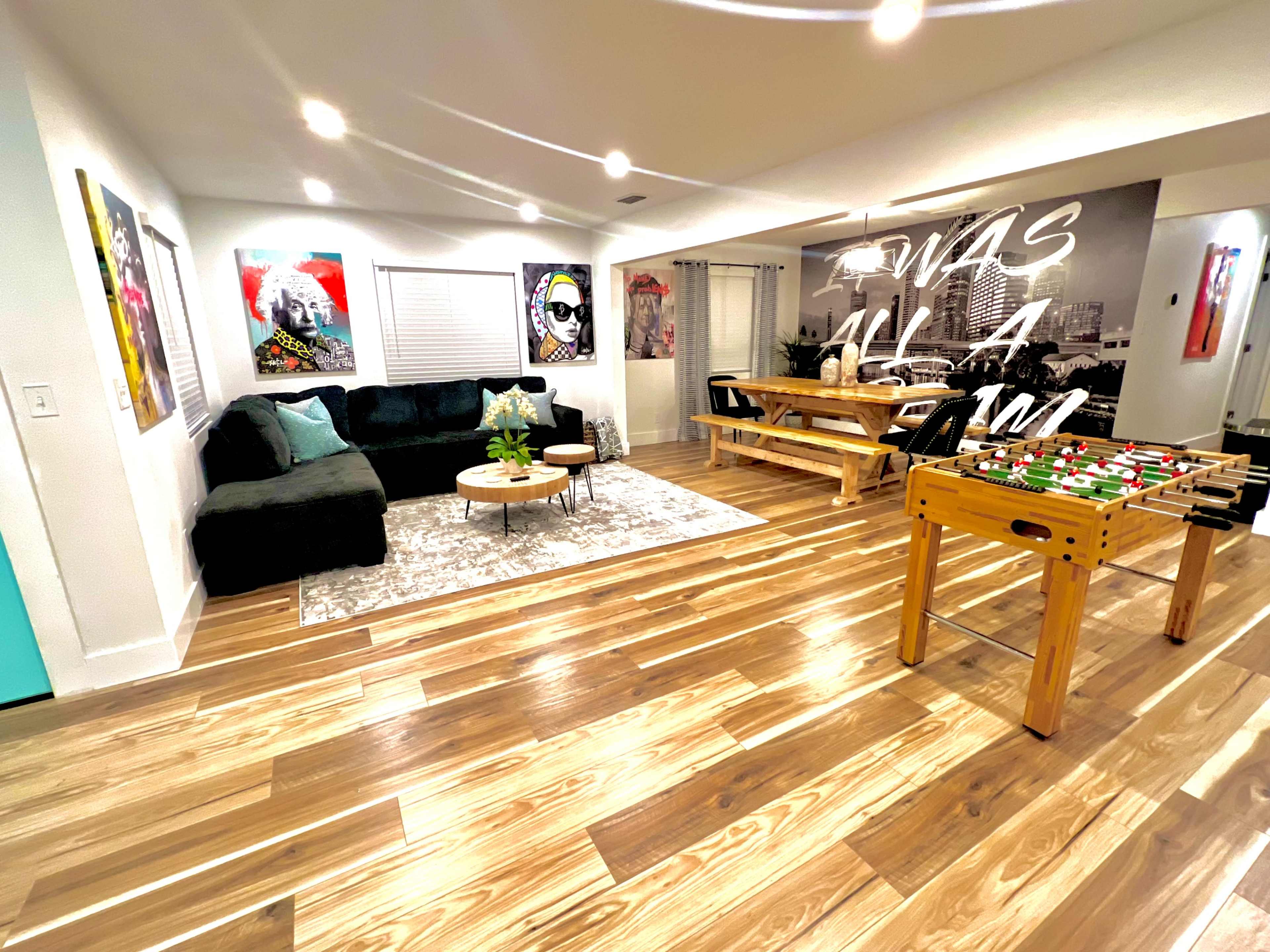 The room features a dark sofa, a round coffee table, a foosball table, and dining area with artwork on the walls and wooden flooring.
