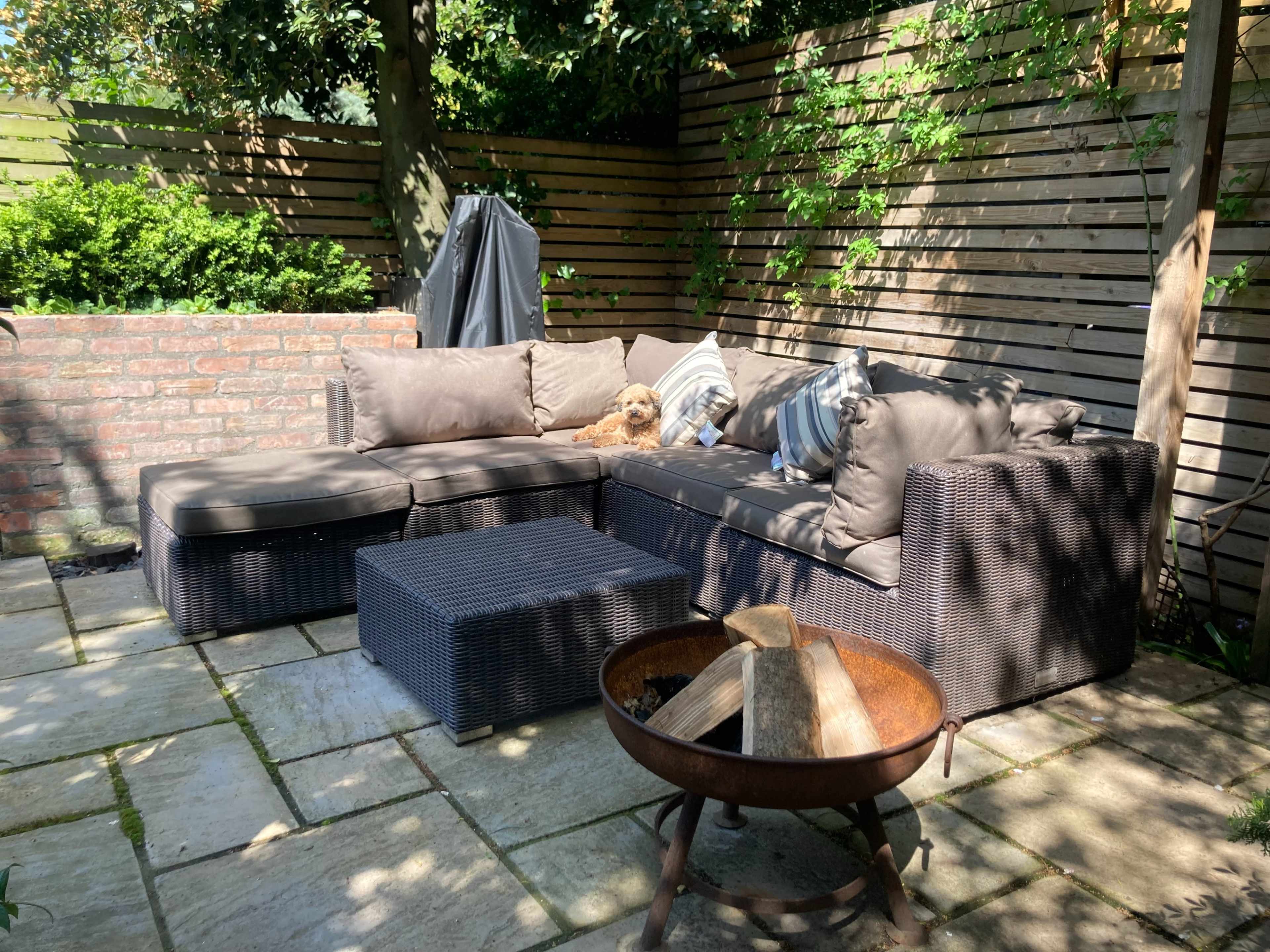 A patio area features a brown wicker sectional sofa with cushions, a matching coffee table, and a metal fire pit surrounded by stone tiles, with a small dog resting on the sofa.