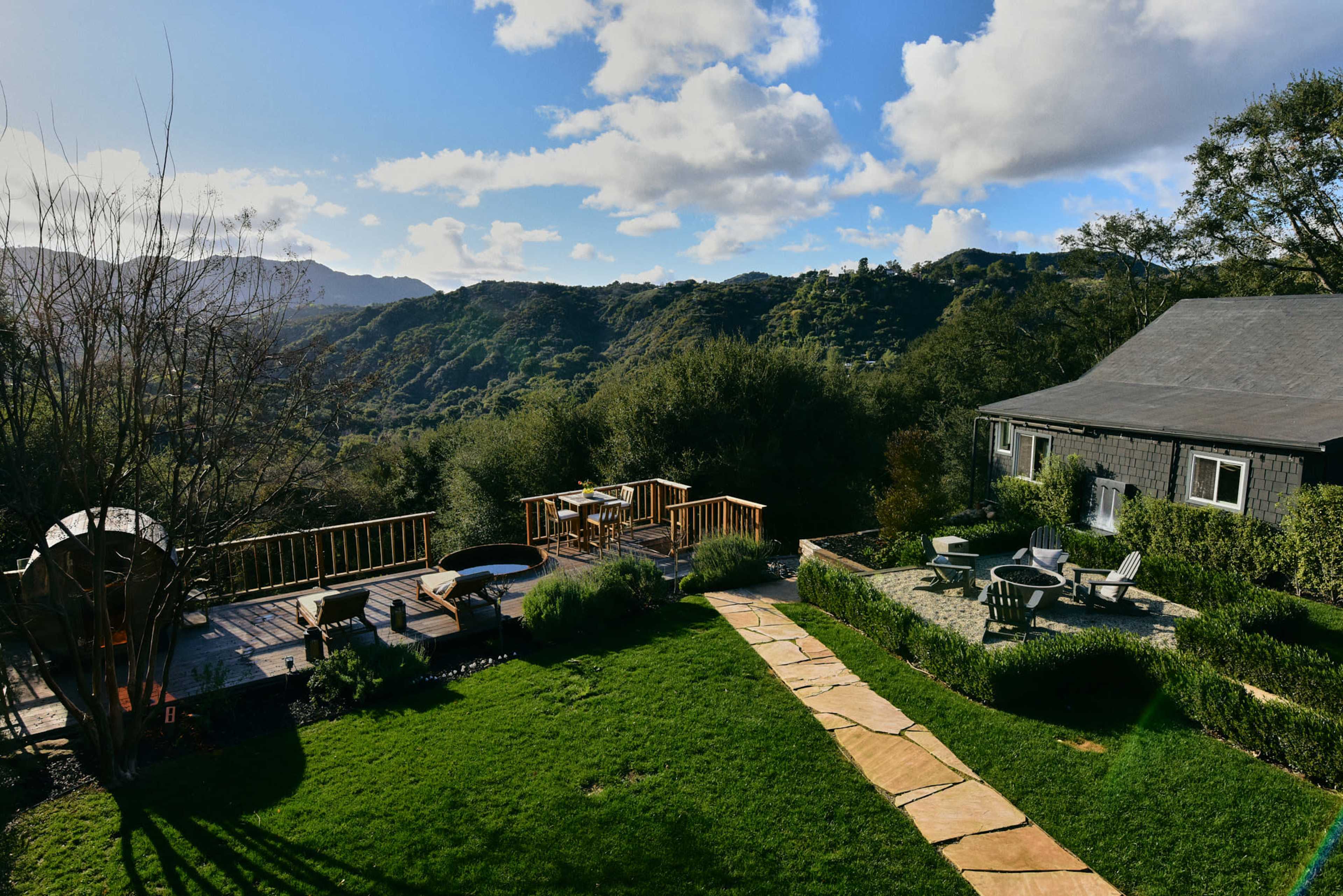 A hillside property features a wooden deck and a stone path surrounded by green grass and trees, with mountains in the background under a partly cloudy sky.