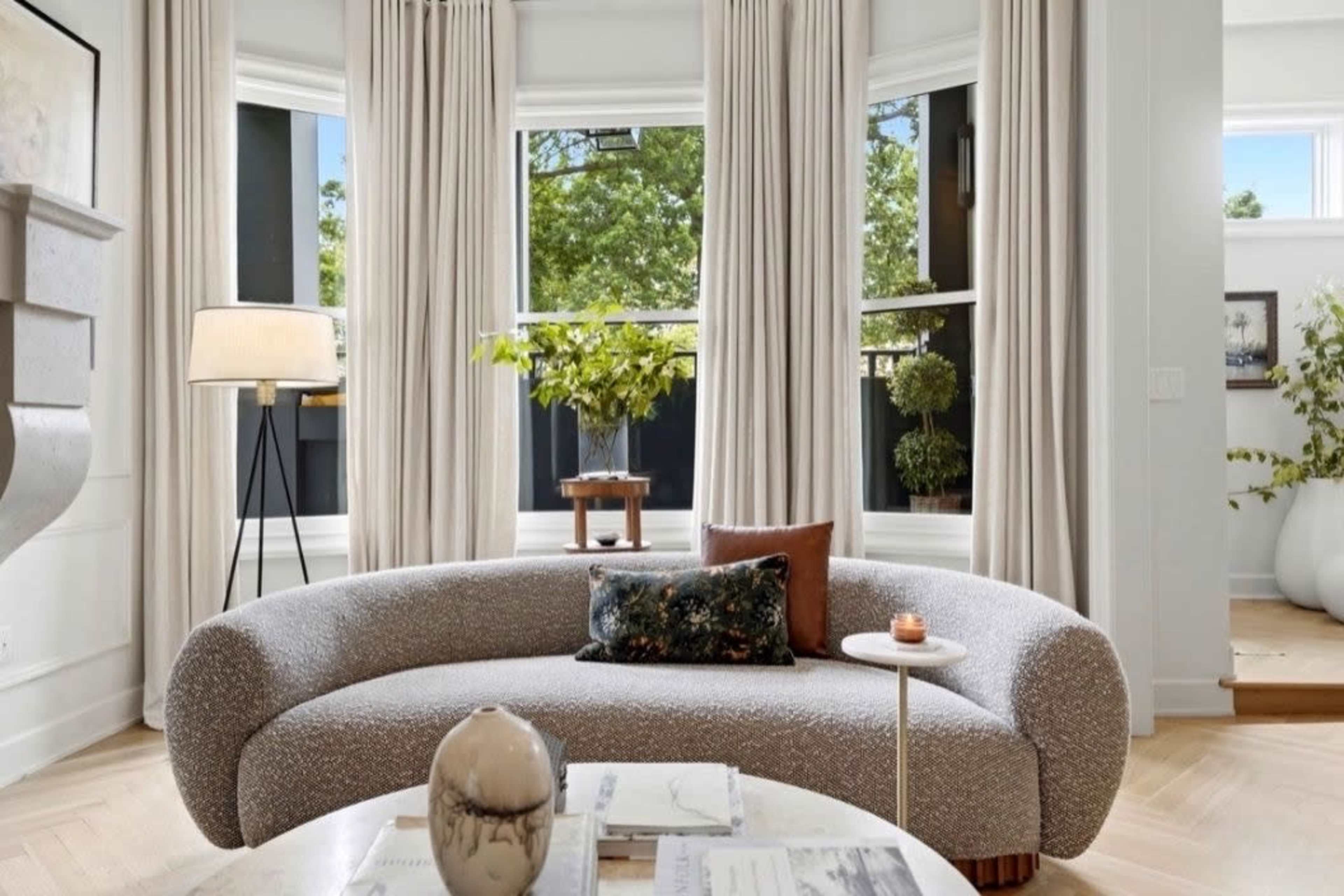 A curved, light-colored sofa is positioned in a bright room with large windows, offering a view of greenery outside.