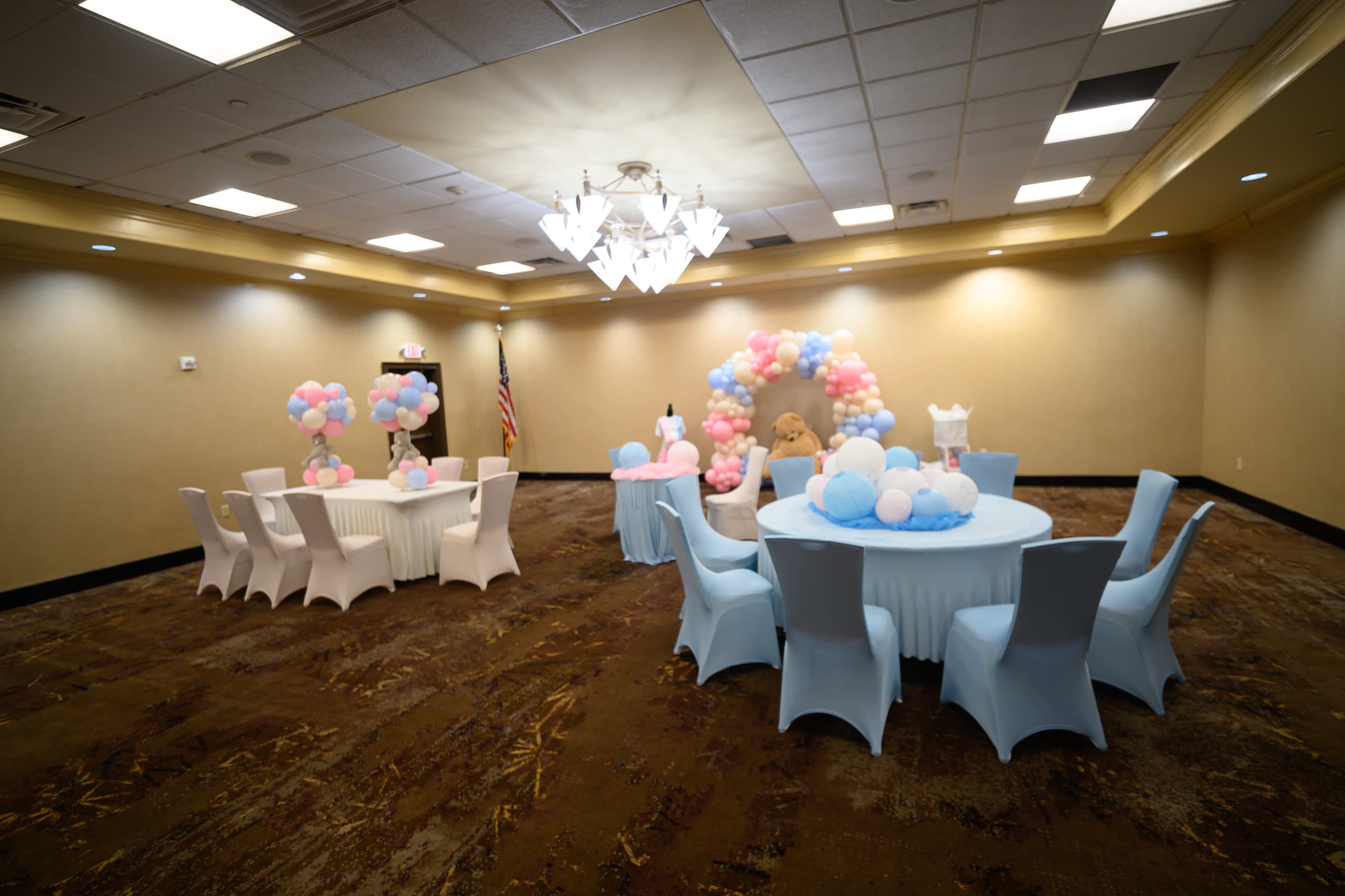 The image shows a decorated event room with tables draped in light blue and white cloths, surrounded by balloon arrangements in shades of pink and blue.