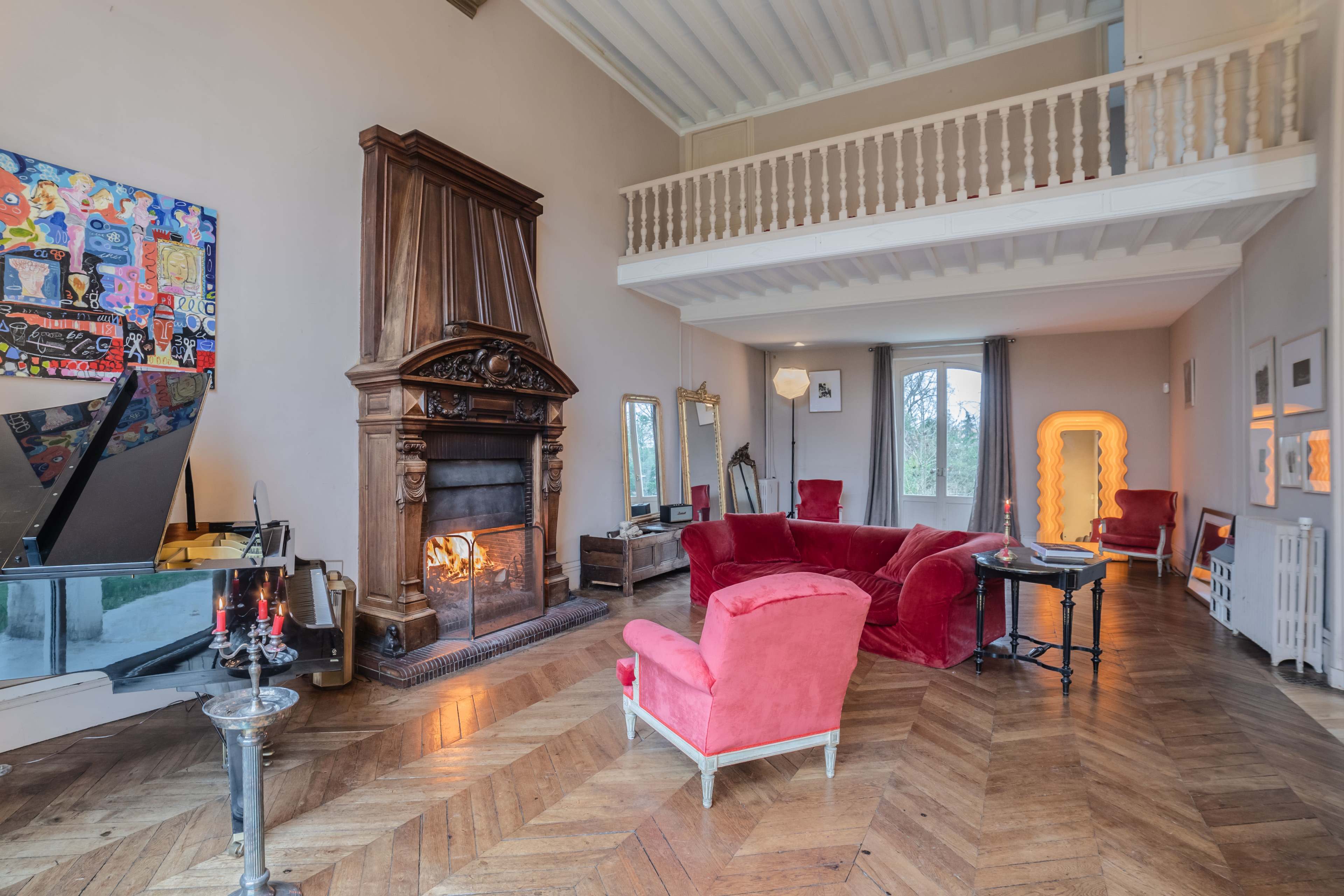 A spacious living room features a large fireplace, a grand piano, and red upholstered seating, with a balcony above and large windows allowing natural light.