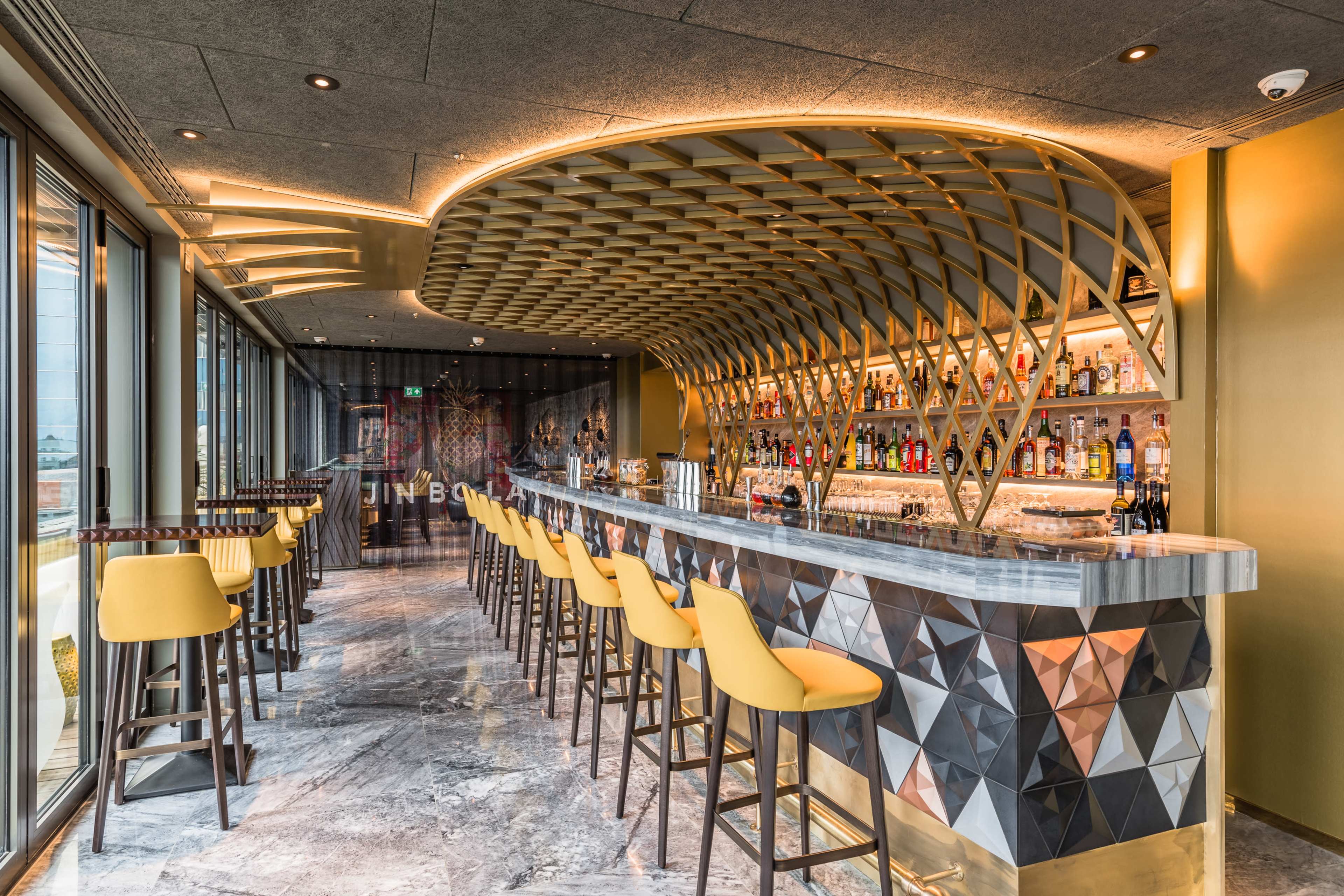 The image shows a modern bar with a curved wooden ceiling, illuminated with warm lighting, and a variety of liquor displayed on shelves behind a geometric-patterned bar counter.
