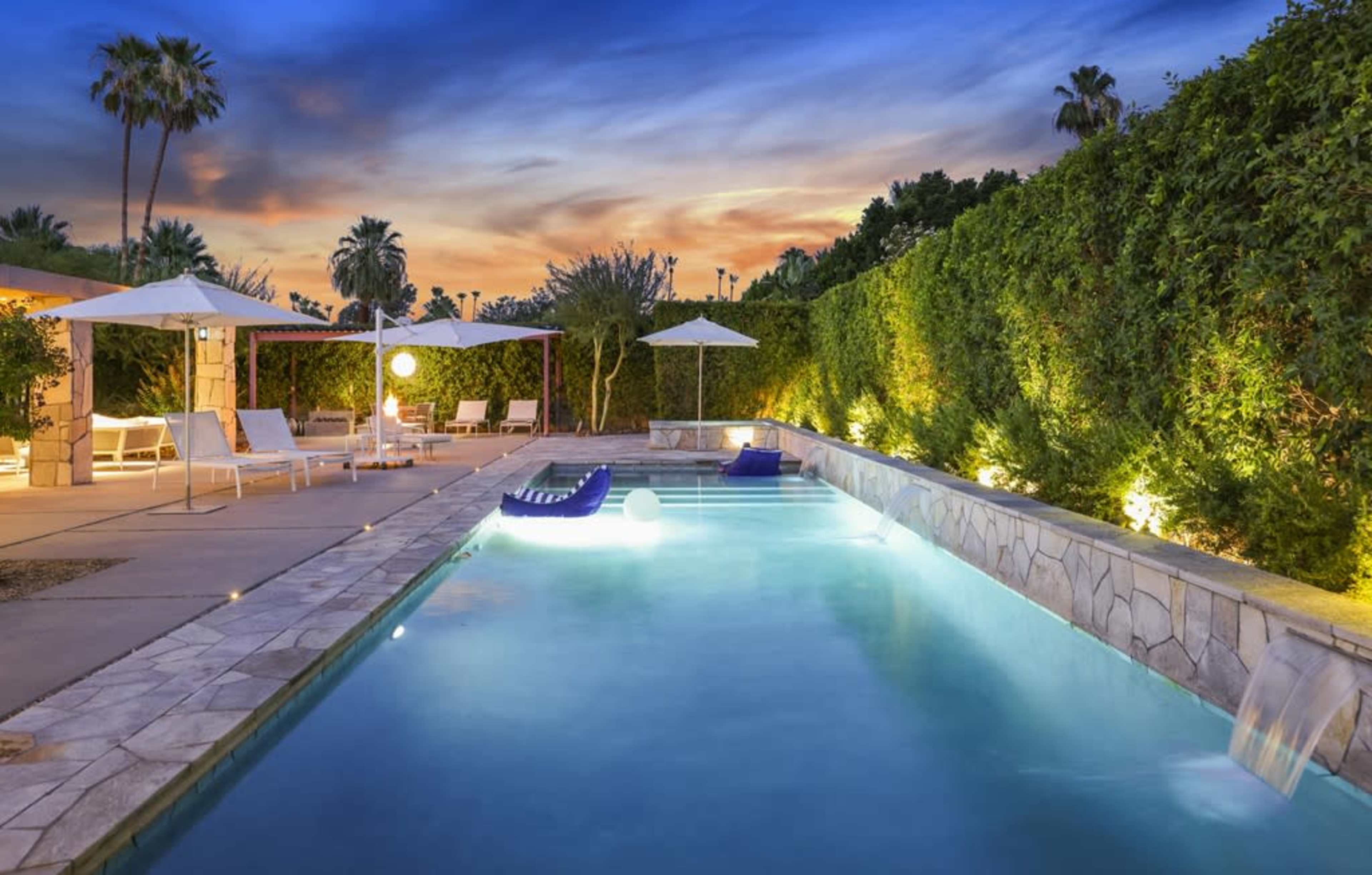 A swimming pool surrounded by lounge chairs and illuminated by landscape lighting is framed by lush greenery and a colorful sunset sky.