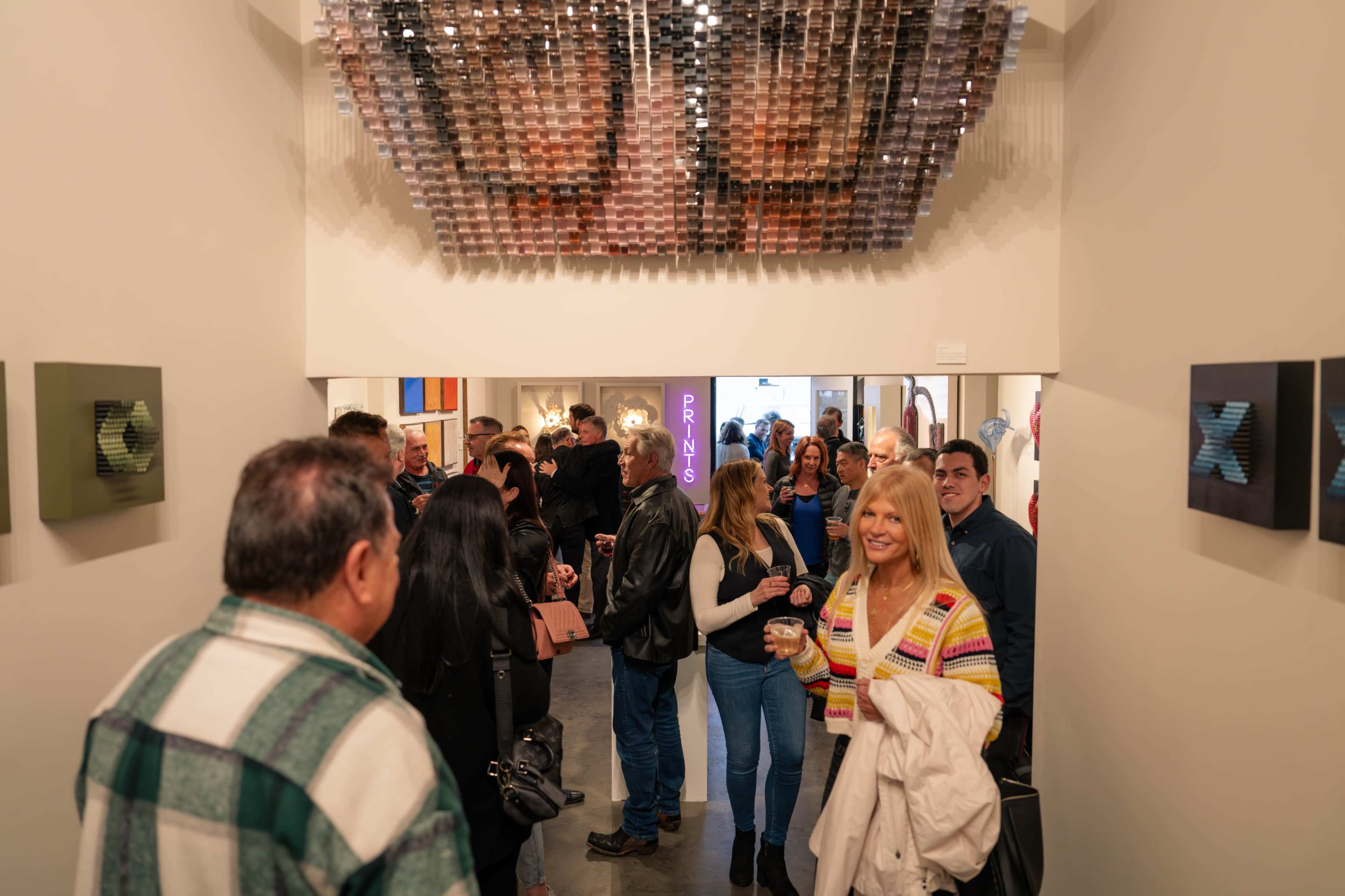 A crowd of people gathers in an art gallery with a reflective installation hanging from the ceiling.
