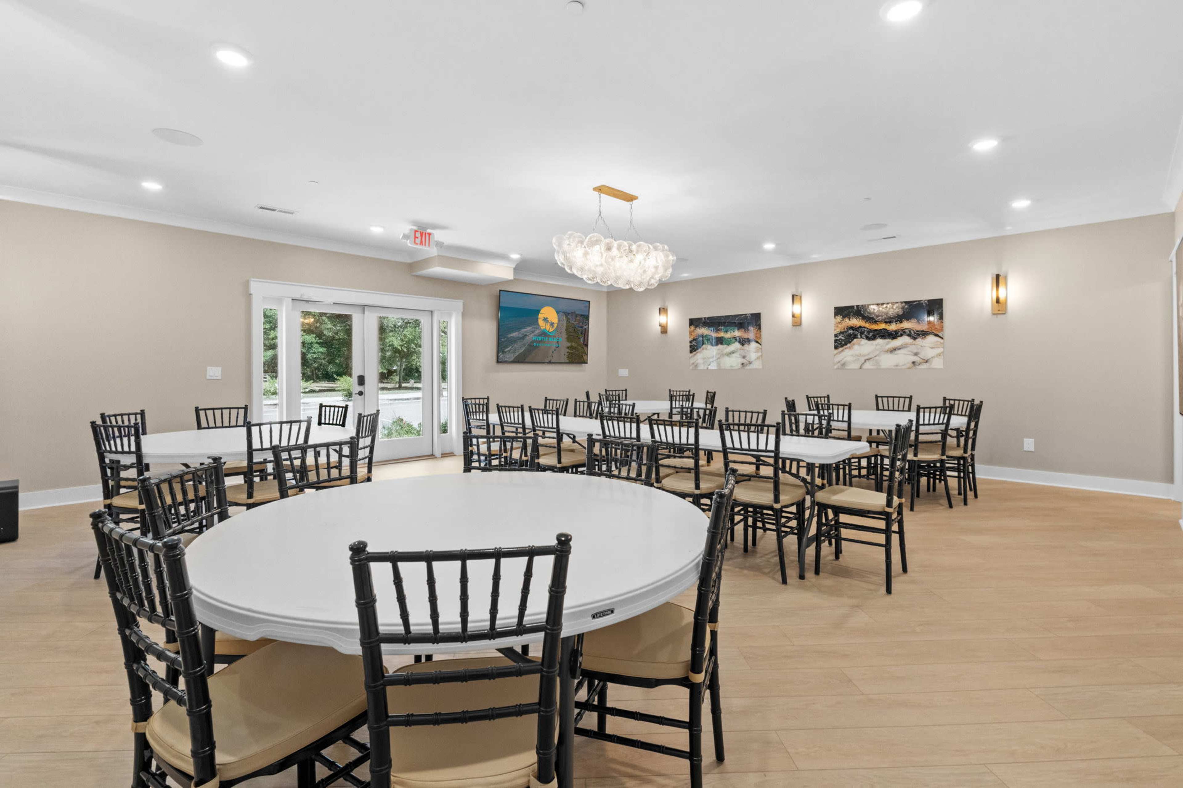 The image shows a well-lit event space with multiple round tables and black chairs, featuring large windows and two decorative artworks on the walls.