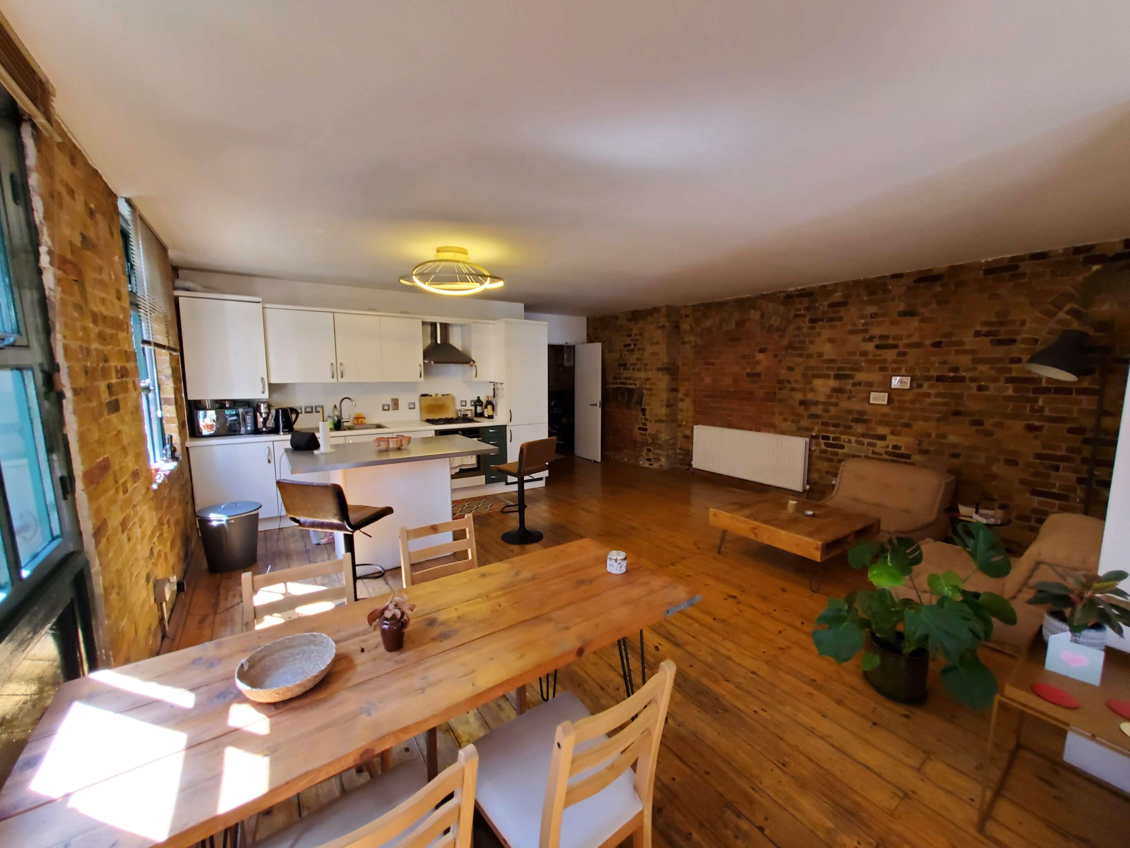 An open-plan living space with a wooden dining table, a kitchen with white cabinets, and a cozy seating area against a brick wall.