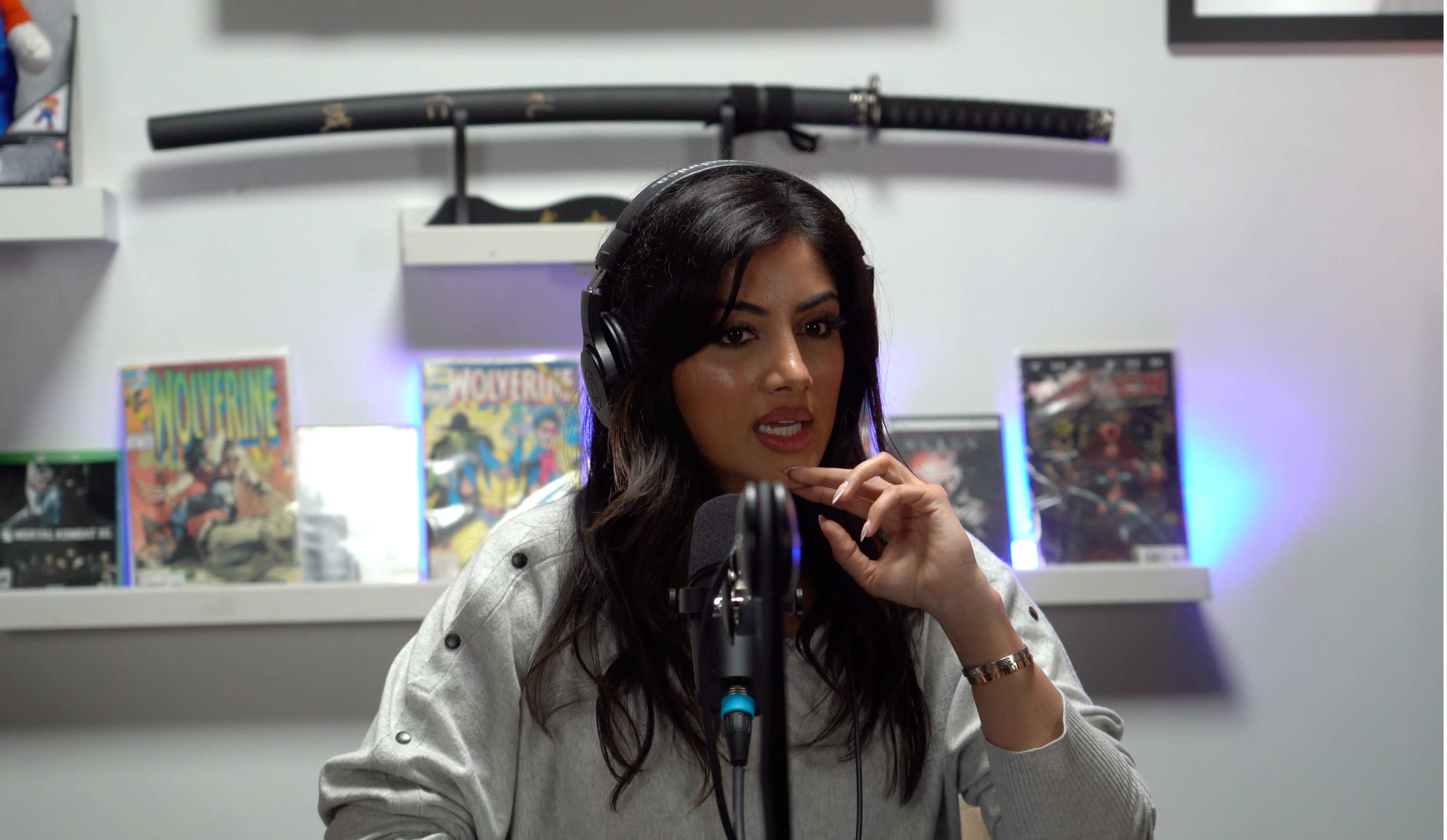 A woman with long dark hair, wearing headphones, is speaking into a microphone in a studio setting with video game and comic-related items displayed on shelves in the background.