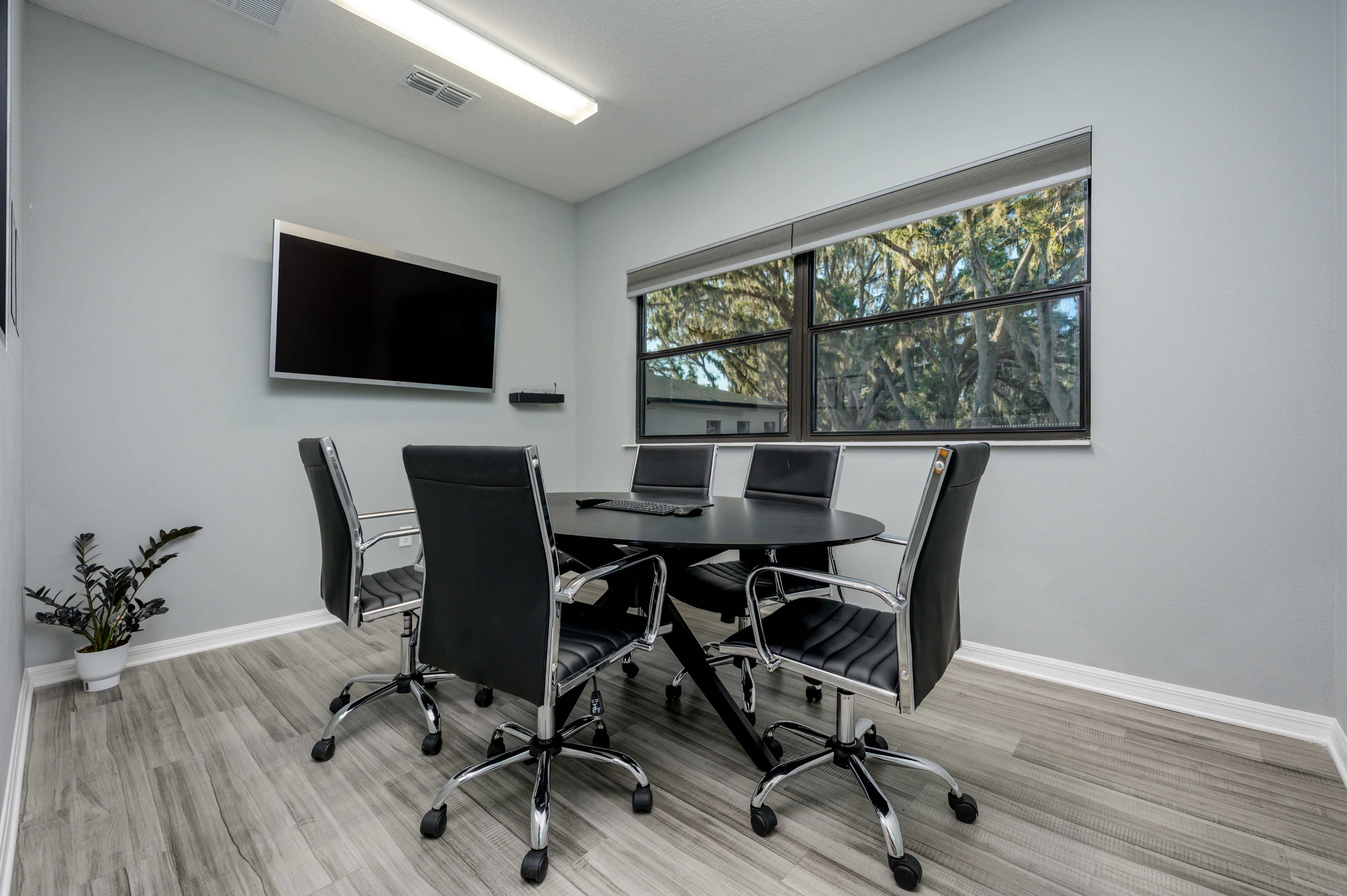 A modern conference room features a round black table surrounded by four black office chairs, with a television mounted on the wall and large windows offering views of trees outside.