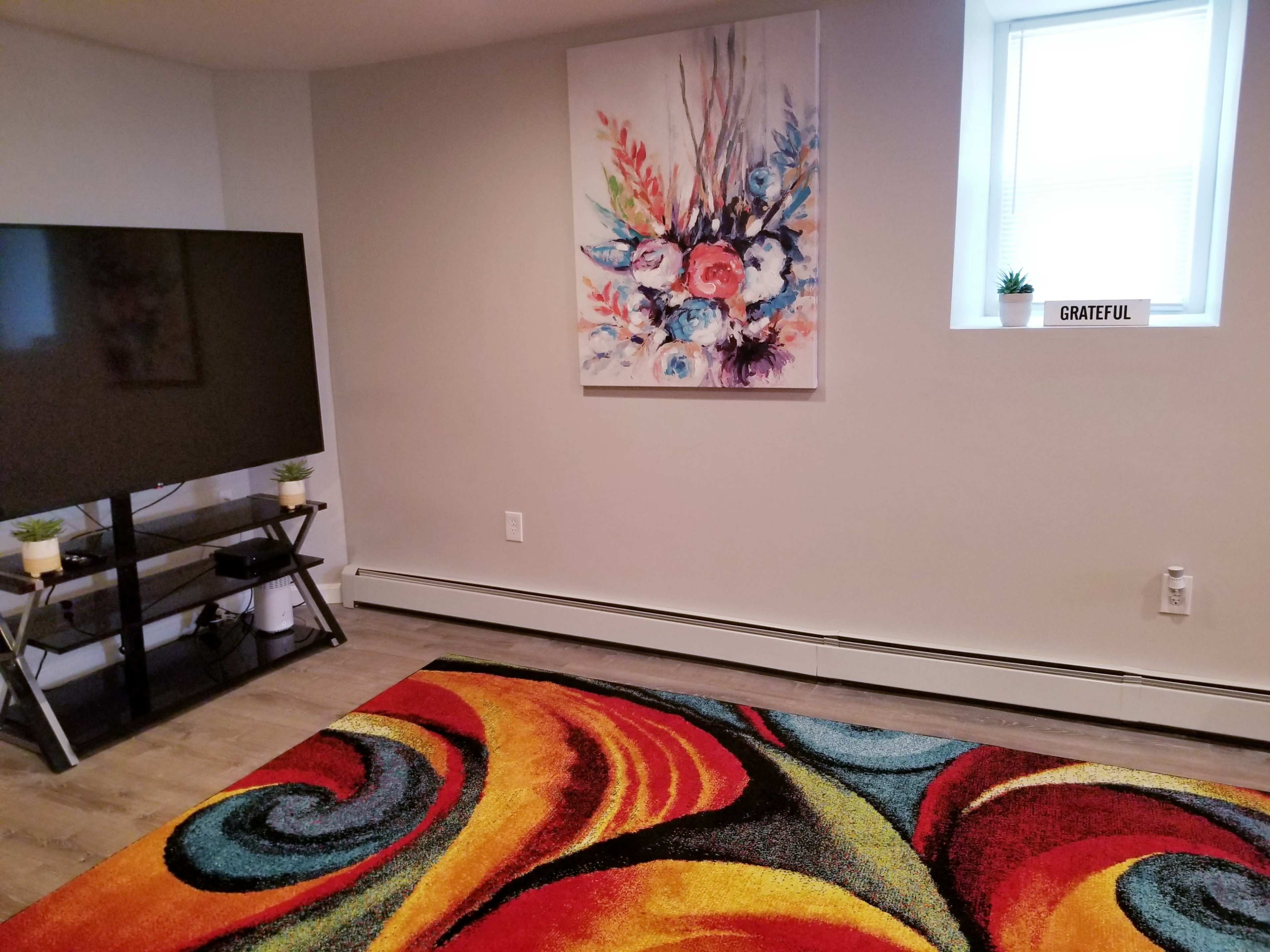 The room features a large television on a stand, a colorful area rug, a floral painting on the wall, and a small window with a sign that reads "GRATEFUL."