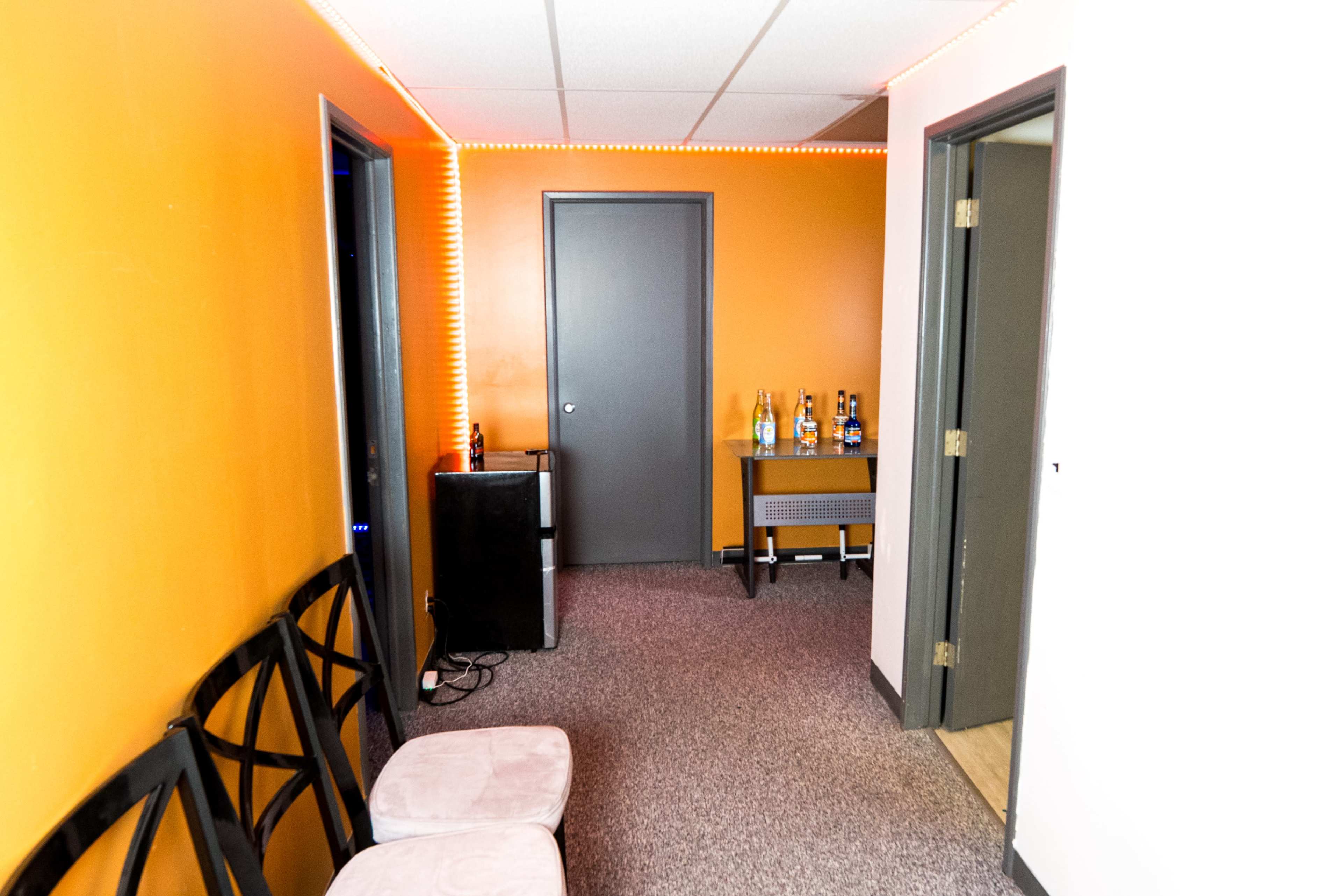 The image shows a narrow hallway with an orange wall, a small table with bottles on it, a black cabinet, and two pink stools.
