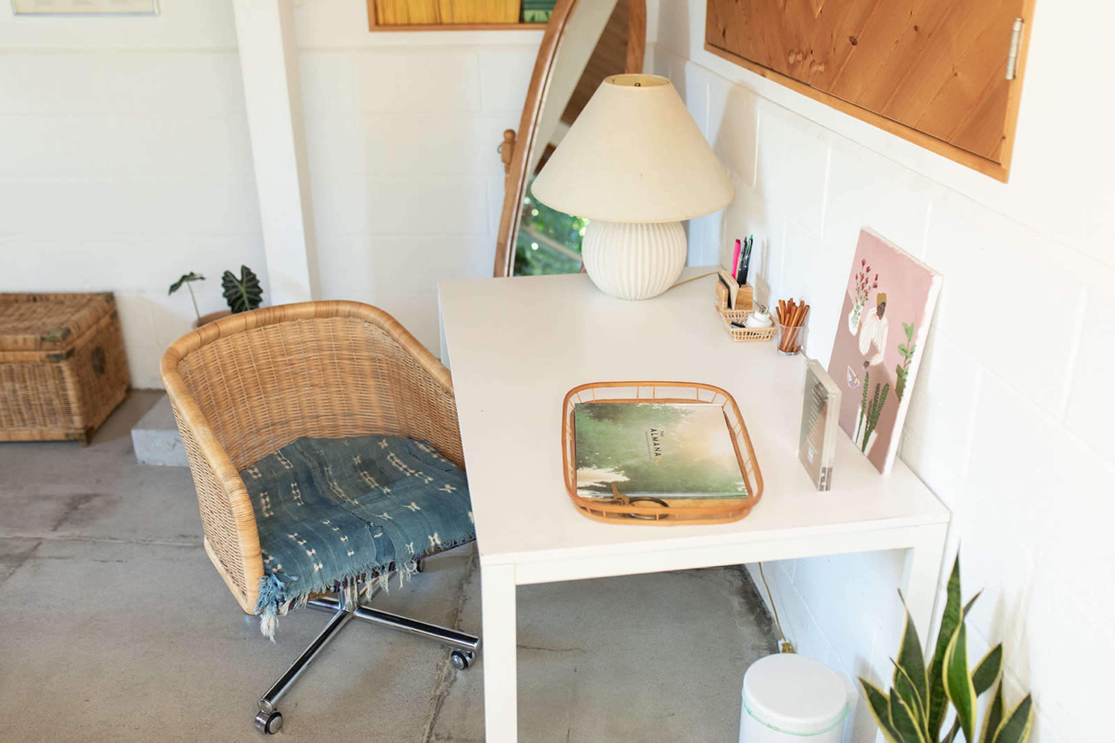 A white desk with a lamp, a tray, and decorative items is accompanied by a wicker chair in a bright, minimalist room.