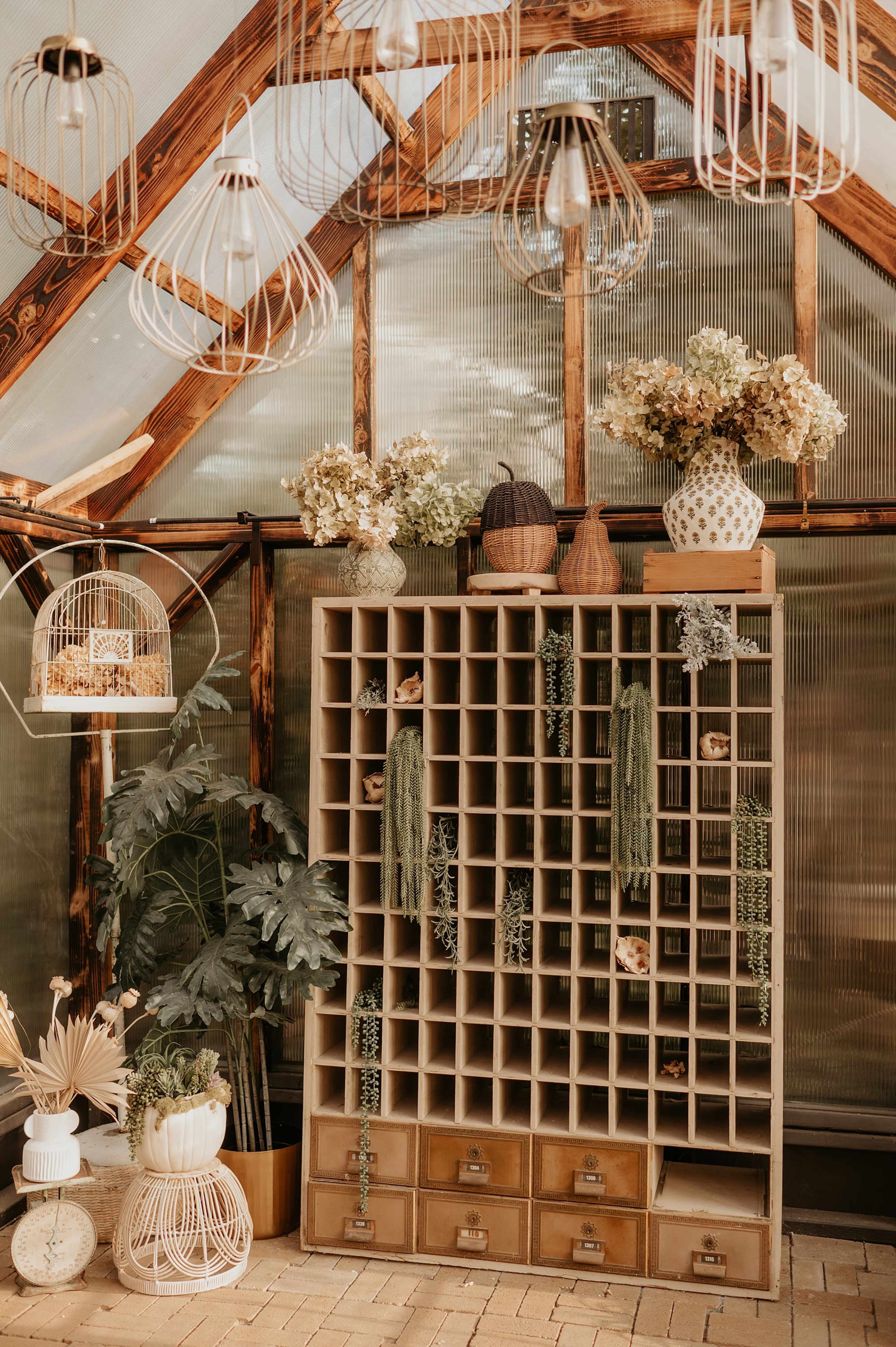 The image shows a neatly organized shelving unit in a greenhouse, adorned with various plants and decorative items, under hanging light fixtures.