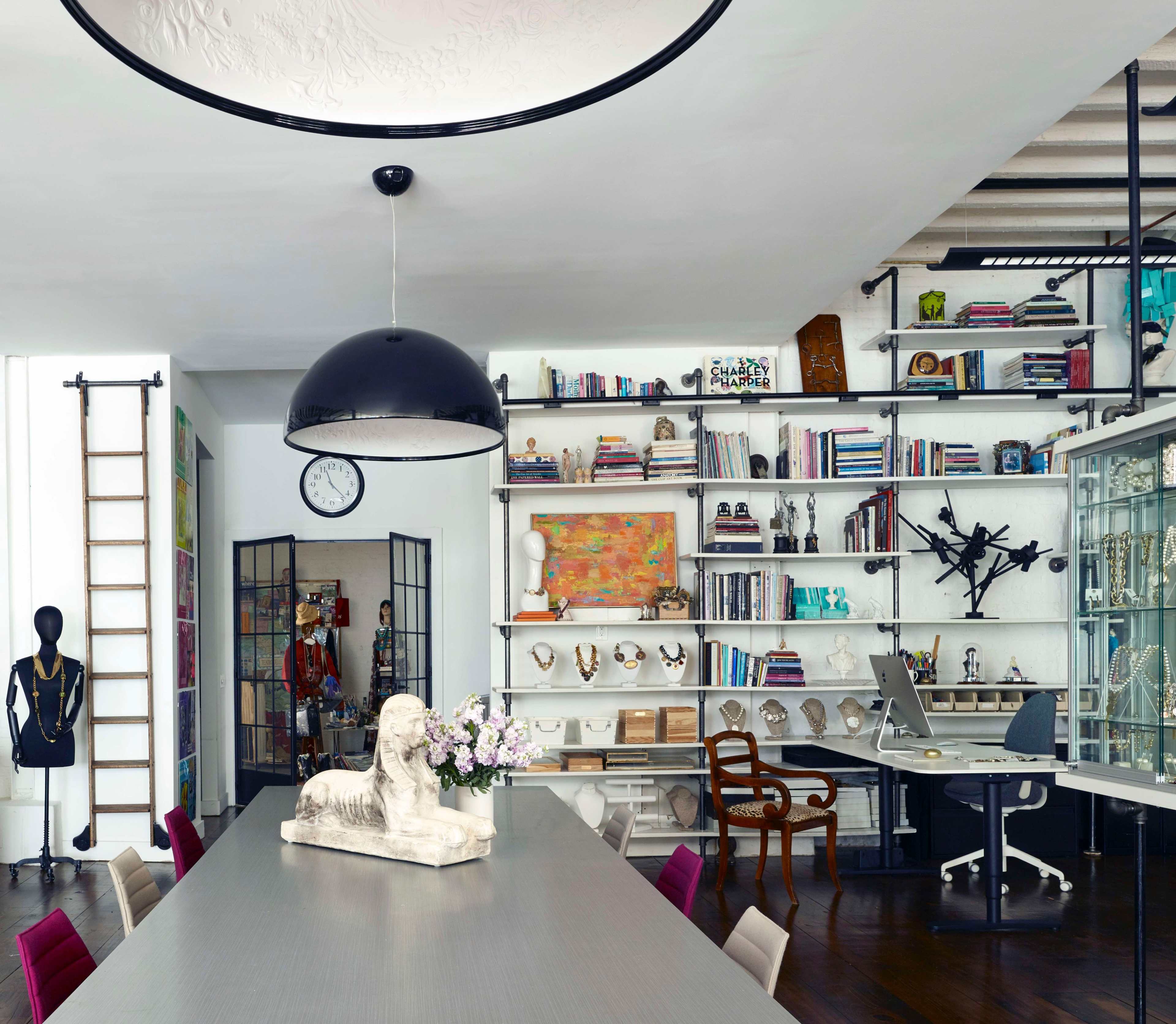 A modern office space features a long table with a sculpture and flowers, shelves lined with books and decorative items, and a workspace with a computer and chair.