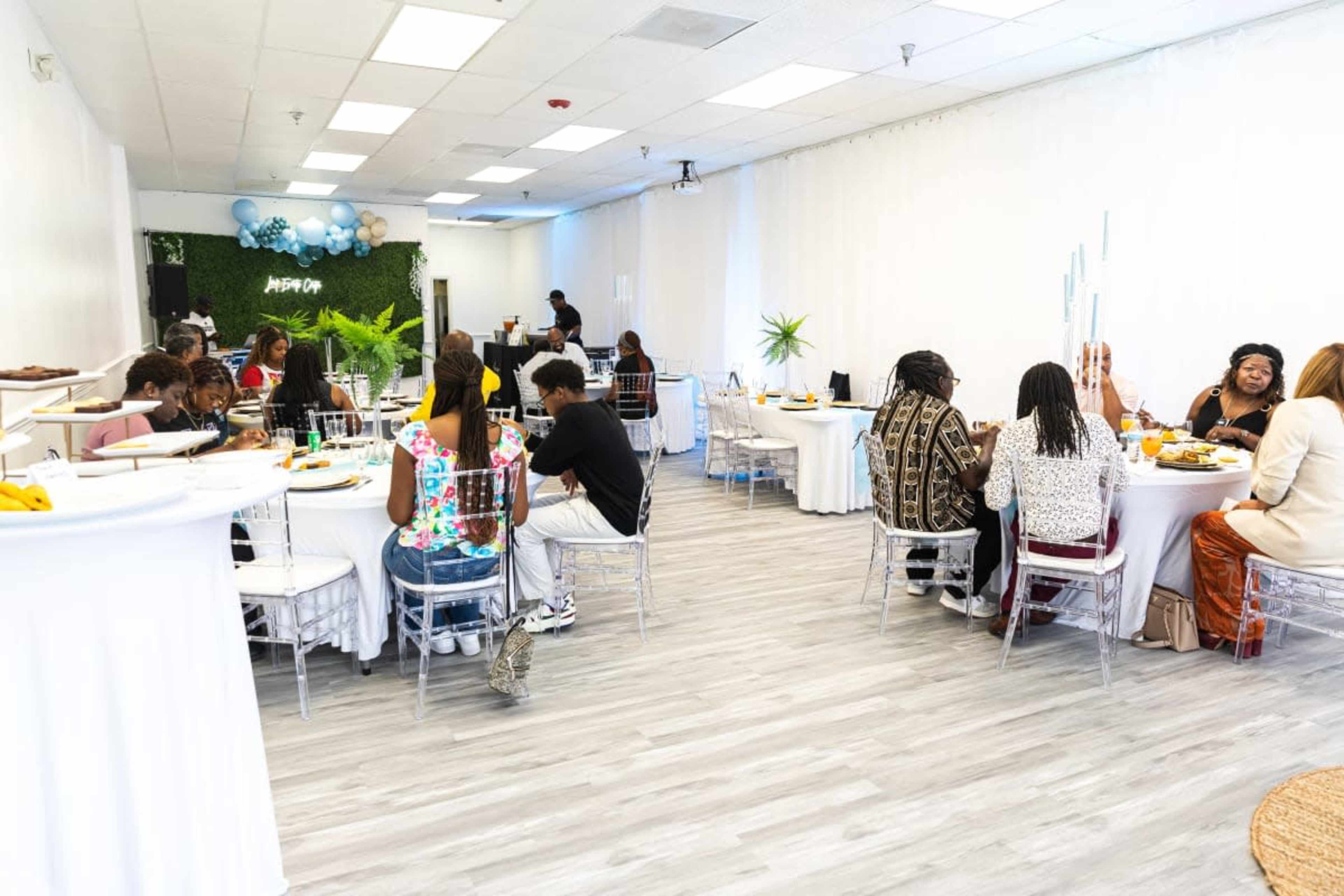 The image shows a modern event space featuring guests seated at round tables enjoying a meal in a bright, airy environment.