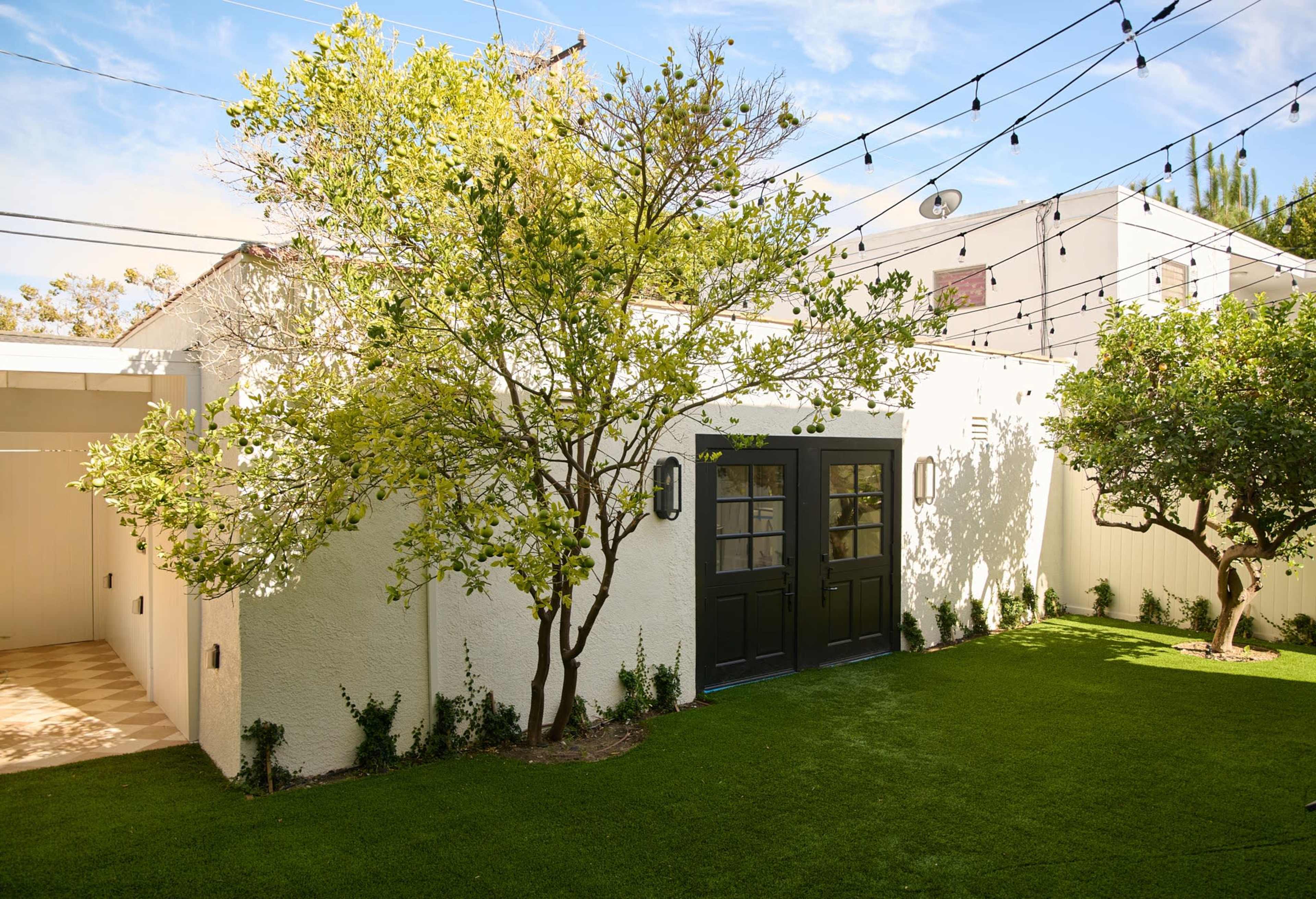 The image depicts a well-maintained backyard with two greenery-laden trees, manicured grass, and a white wall featuring black double doors.