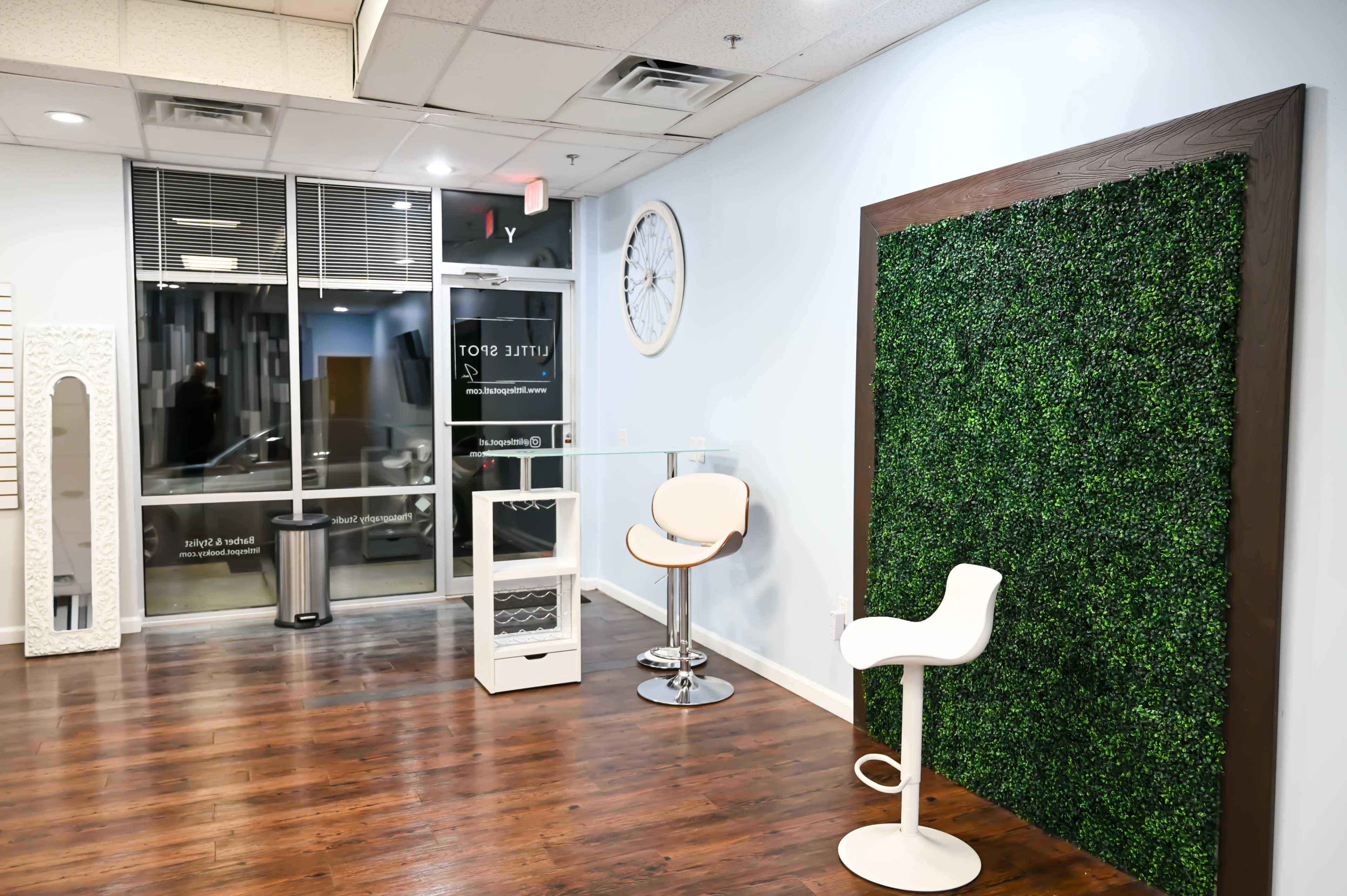 A minimalist interior space featuring a green moss wall, a glass table with two stools, and large windows with light coming in.