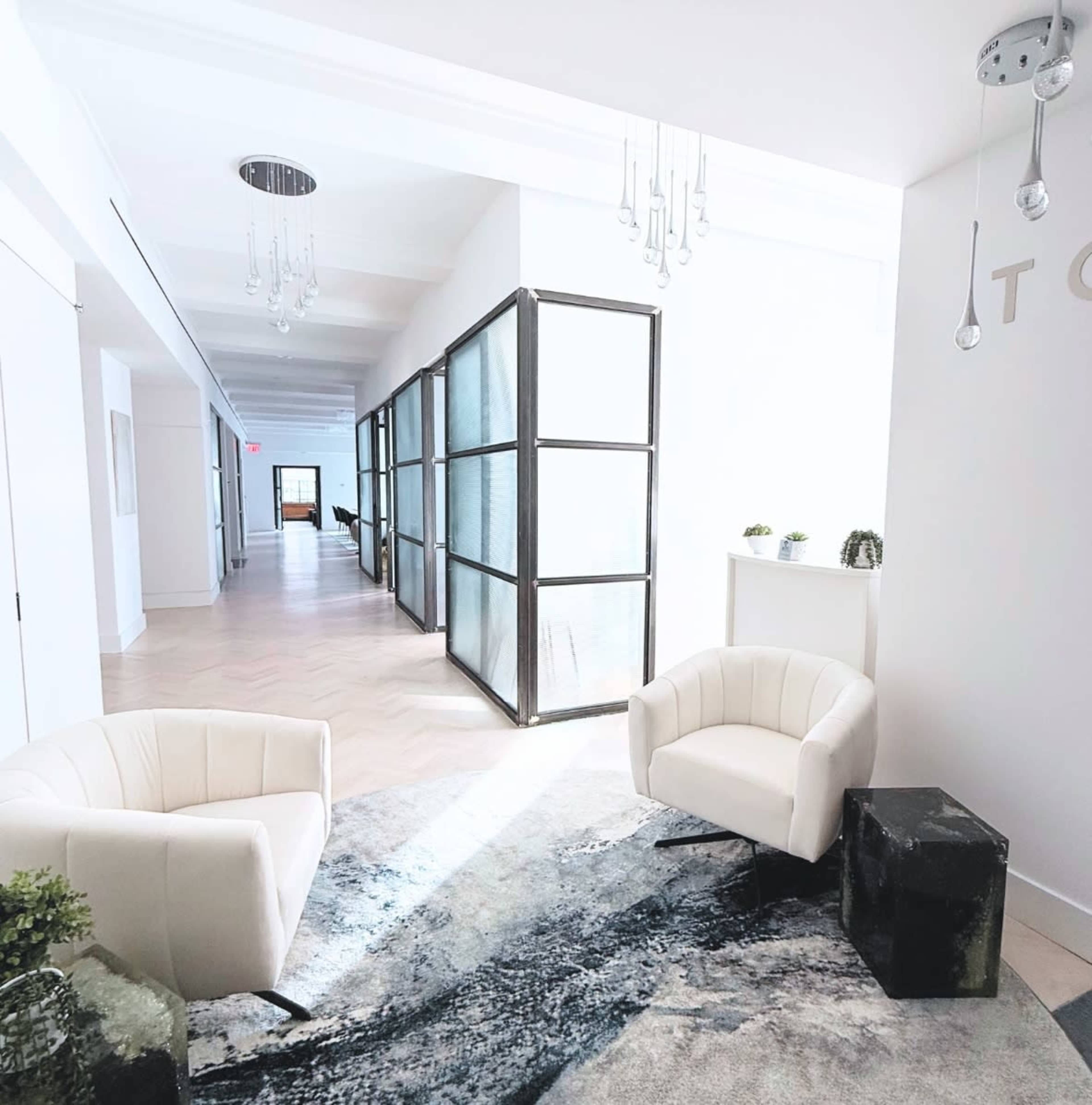 The image shows a well-lit hallway featuring two white armchairs, a dark cube side table, and a partially frosted glass partition, with light streaming in from large windows.