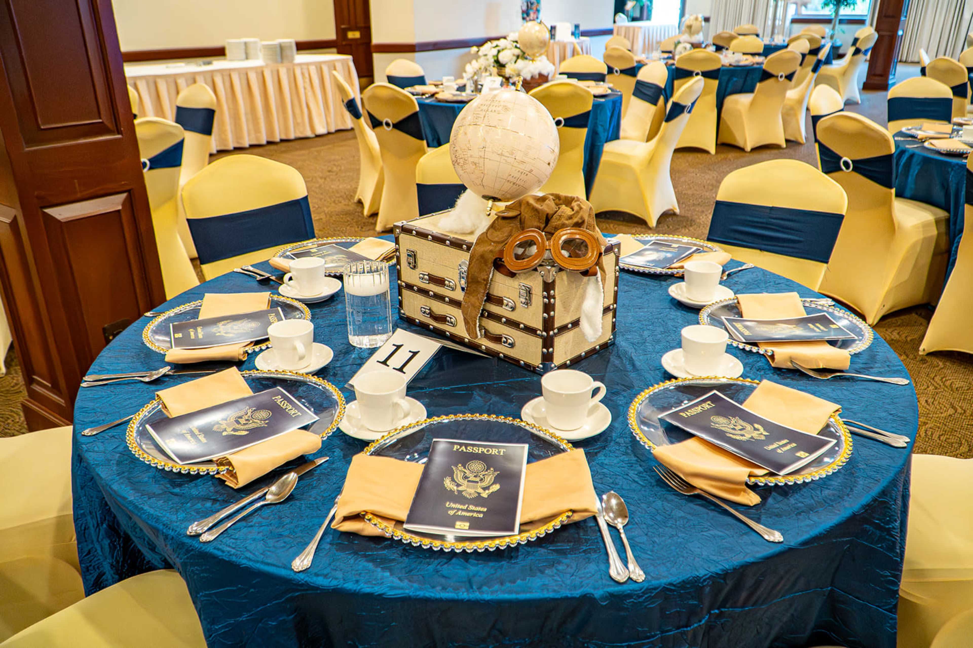 The image shows a elegantly set table with a blue tablecloth, yellow chair covers, and decorative items, including a vintage-style suitcase, glasses, and menus titled "PASSPORT."