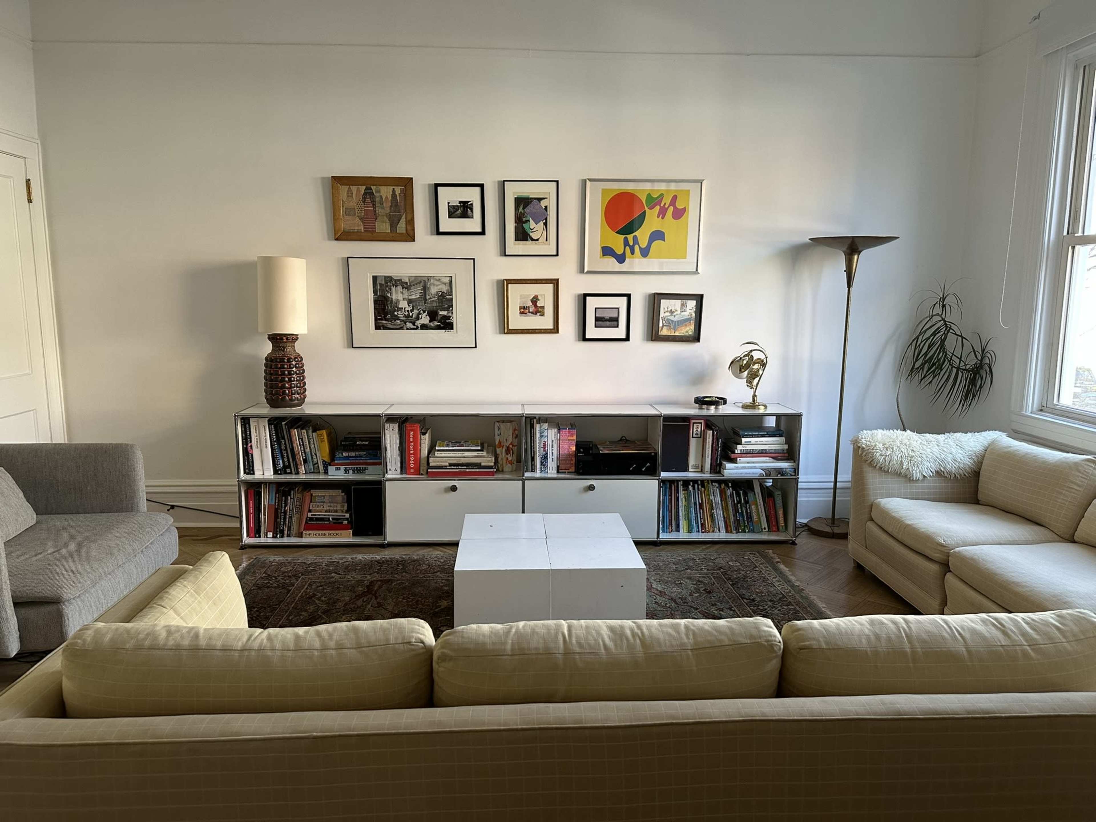 The image shows a cozy living room with a light-colored sofa facing a low white coffee table, surrounded by bookshelves filled with books and decorative items, and adorned with framed artwork on the wall.