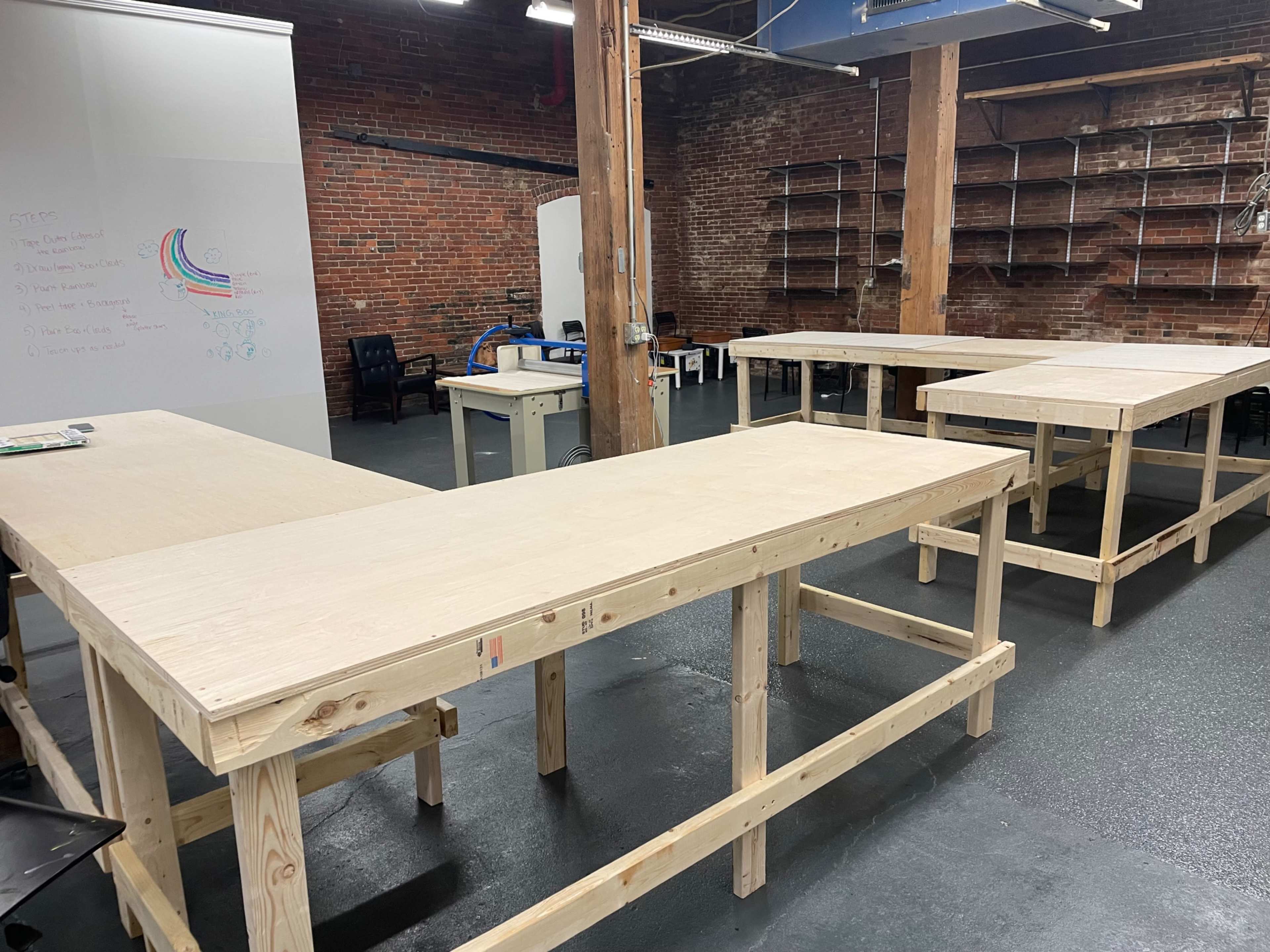 A spacious room with exposed brick walls, featuring multiple wooden work tables arranged in a U-shape on a smooth floor.